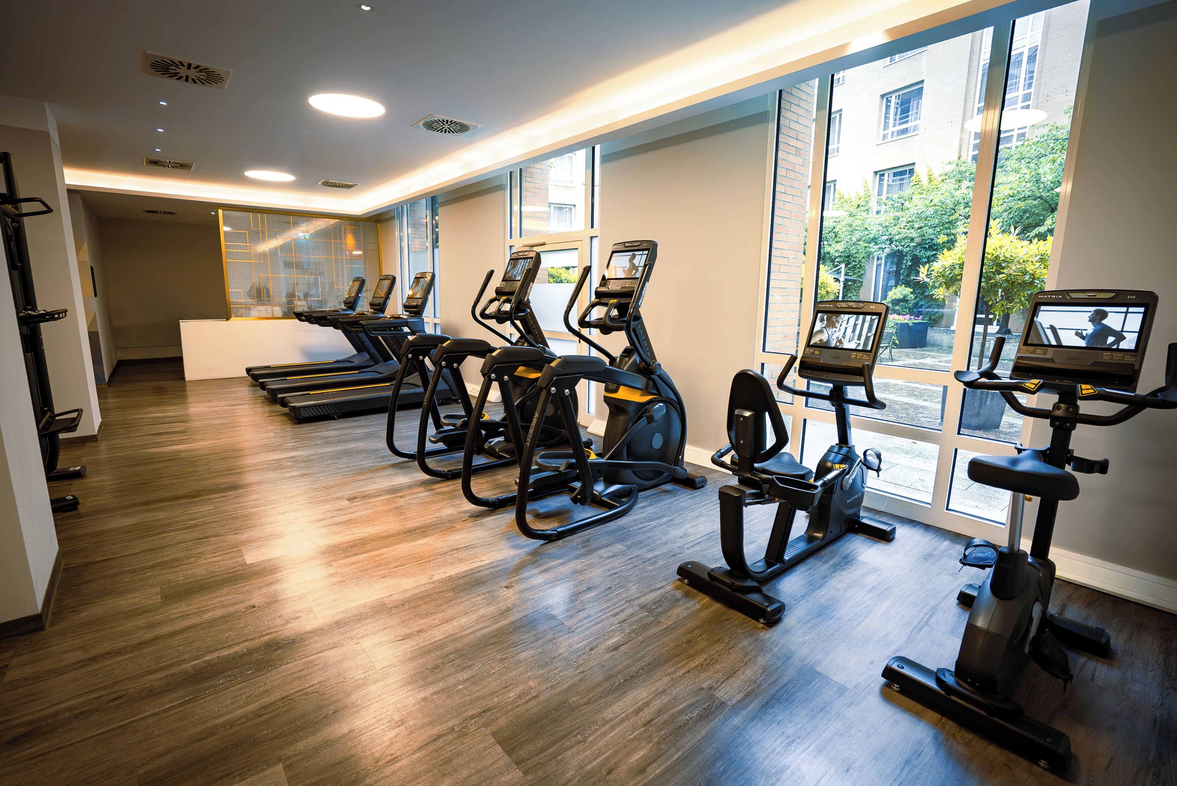 Un gimnasio con cintas de correr y máquinas elípticas, ventanas amplias y suelo de madera.