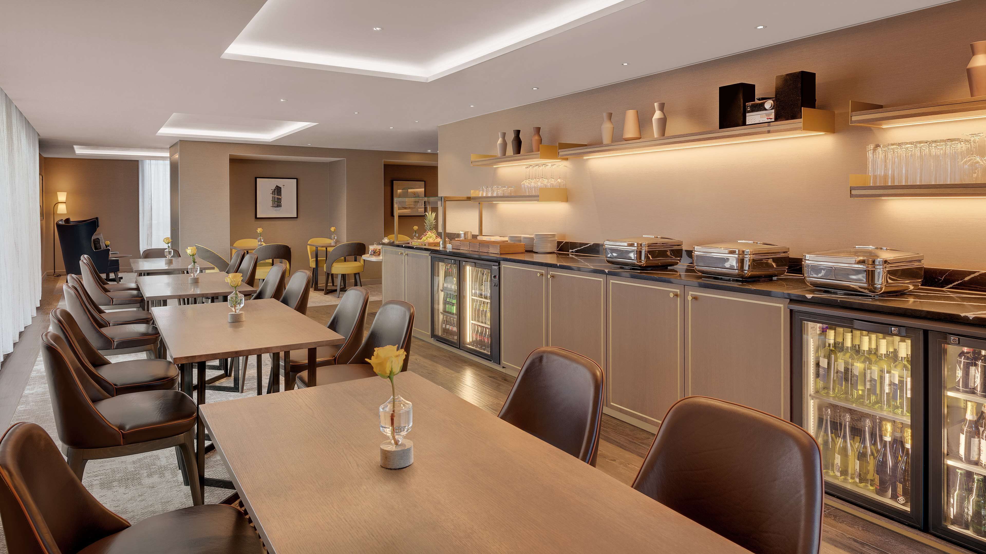 A modern dining area with long tables and chairs, a bar with glassware, and a refrigerator stocked with beverages.