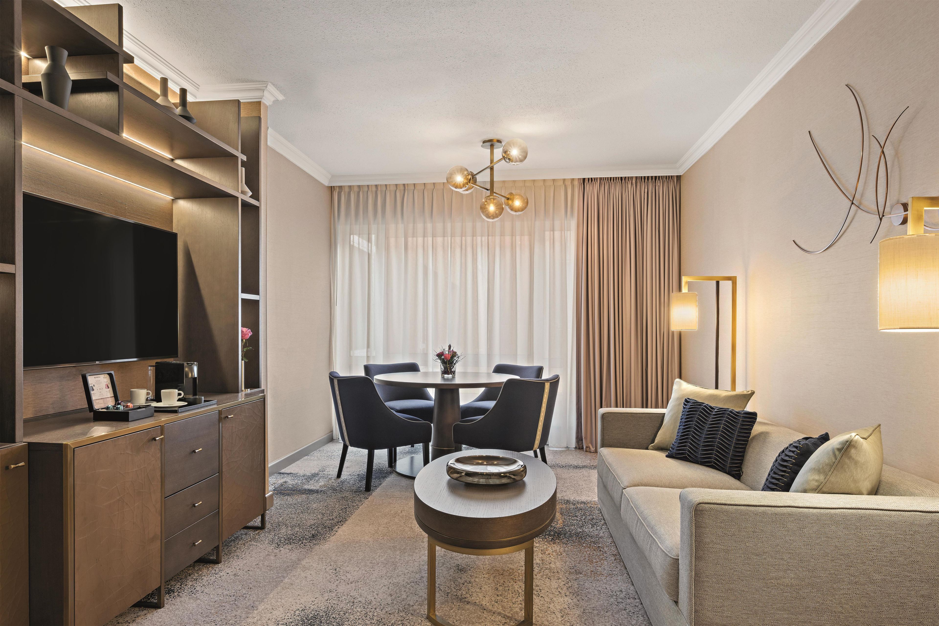 A modern hotel room with a flat-screen TV, a dining table with chairs, a sofa, and a coffee table.