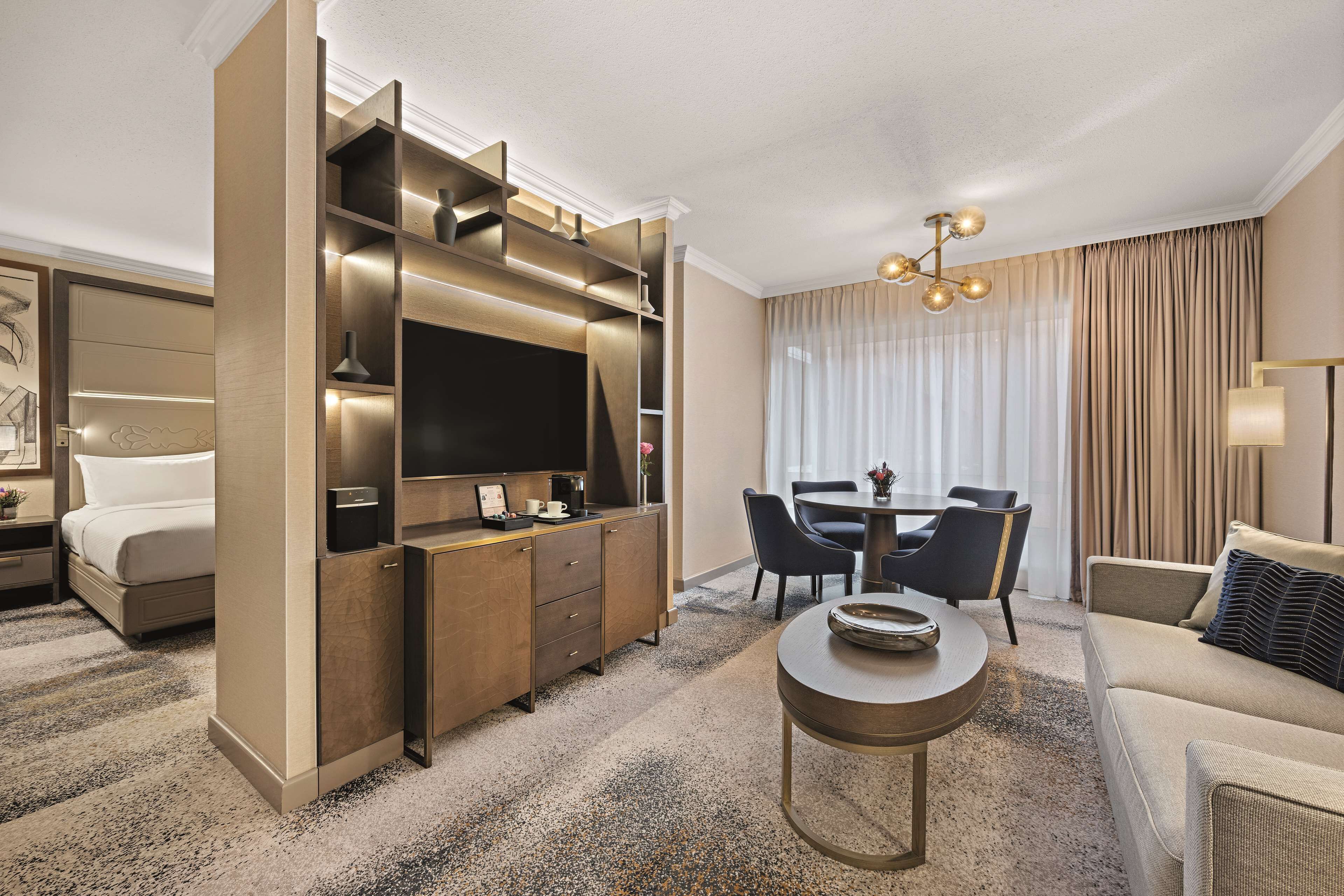 Una habitación de hotel moderna con cama, escritorio con televisor, zona de comedor con sillas y sofá.
