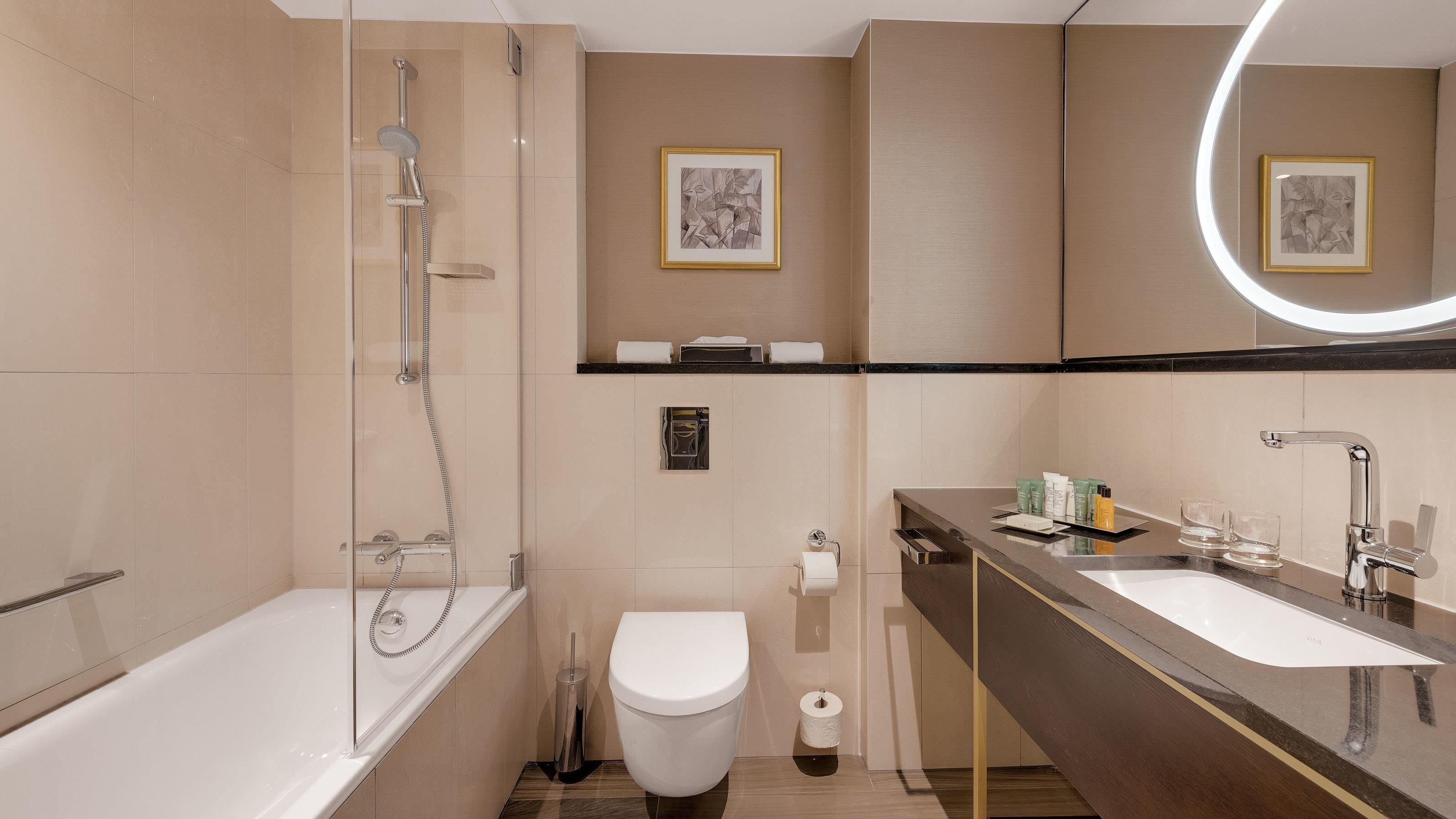 A modern bathroom with a glass-enclosed shower, a toilet, a sink with toiletries, and framed artwork on the wall.