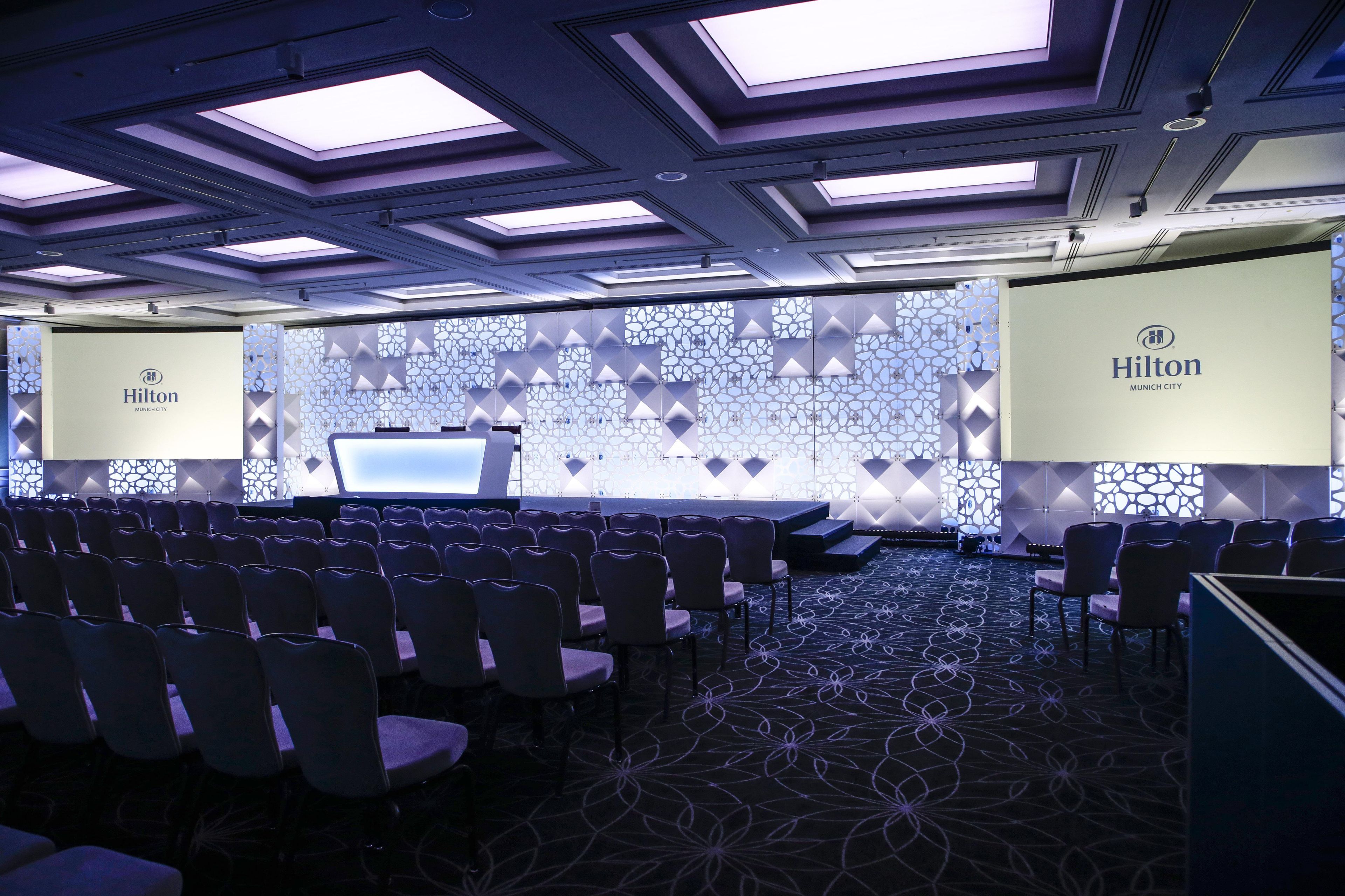 A conference room with rows of chairs, a presentation screen, and a Hilton logo on the wall.