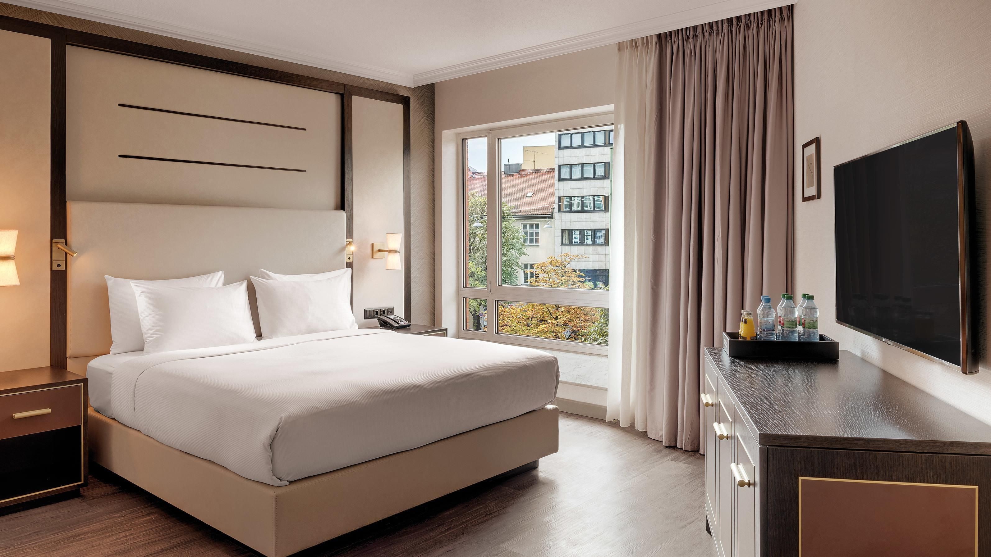 A hotel room with a large bed, a desk with a flat-screen TV, a window with curtains, and a view of buildings outside.