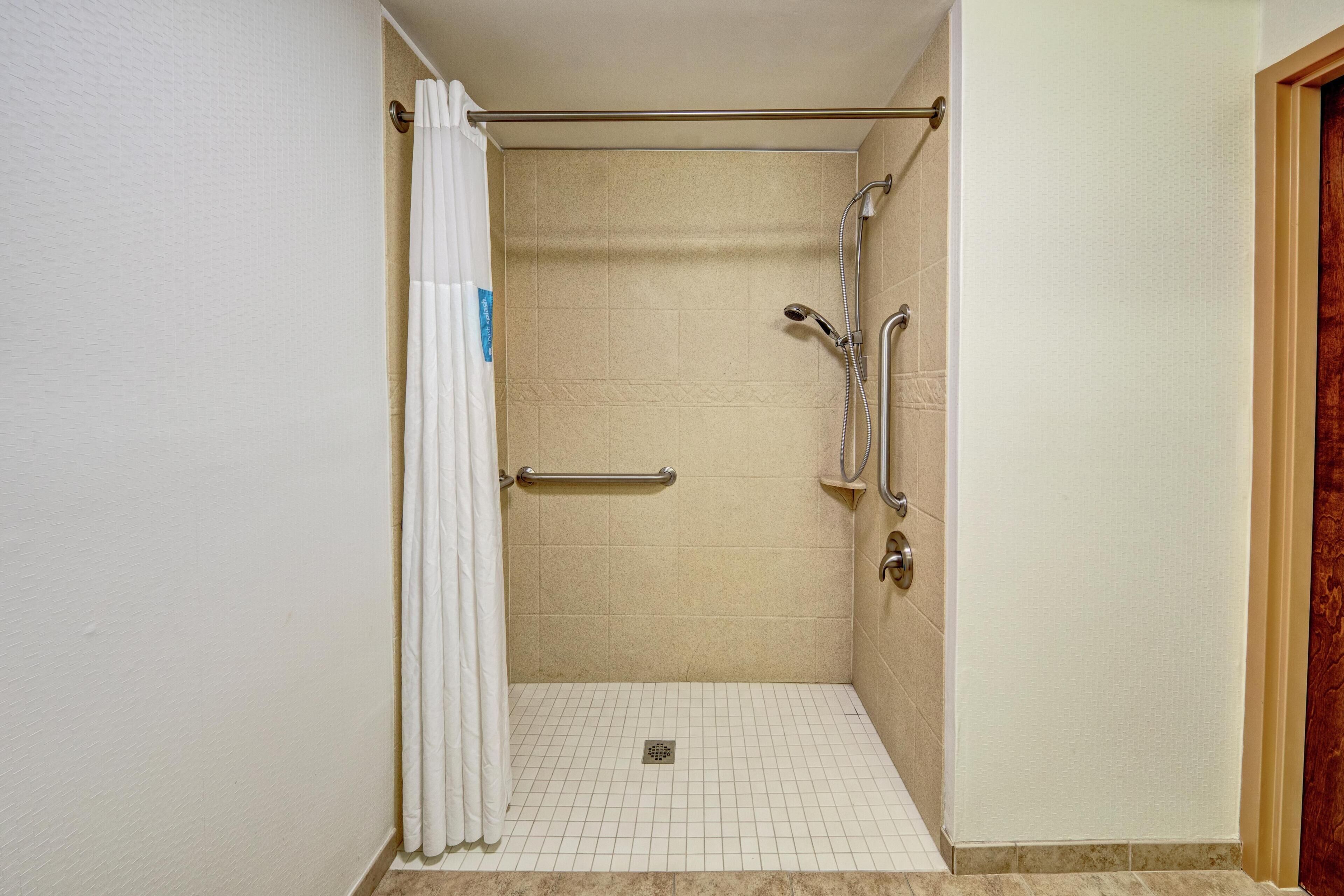 A wheelchair-accessible shower stall with a white curtain, grab bars, and a tiled floor.