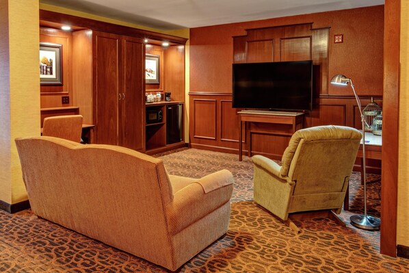 Habitación de hotel con sofá, sillón, televisión, kitchenette y alfombra estampada.