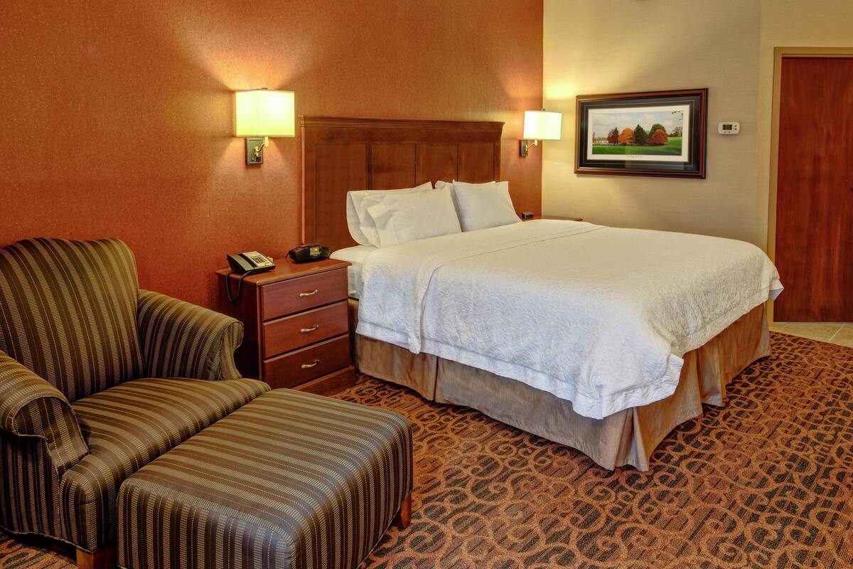 A hotel room with a bed, a chair, a nightstand, and a wall-mounted lamp.
