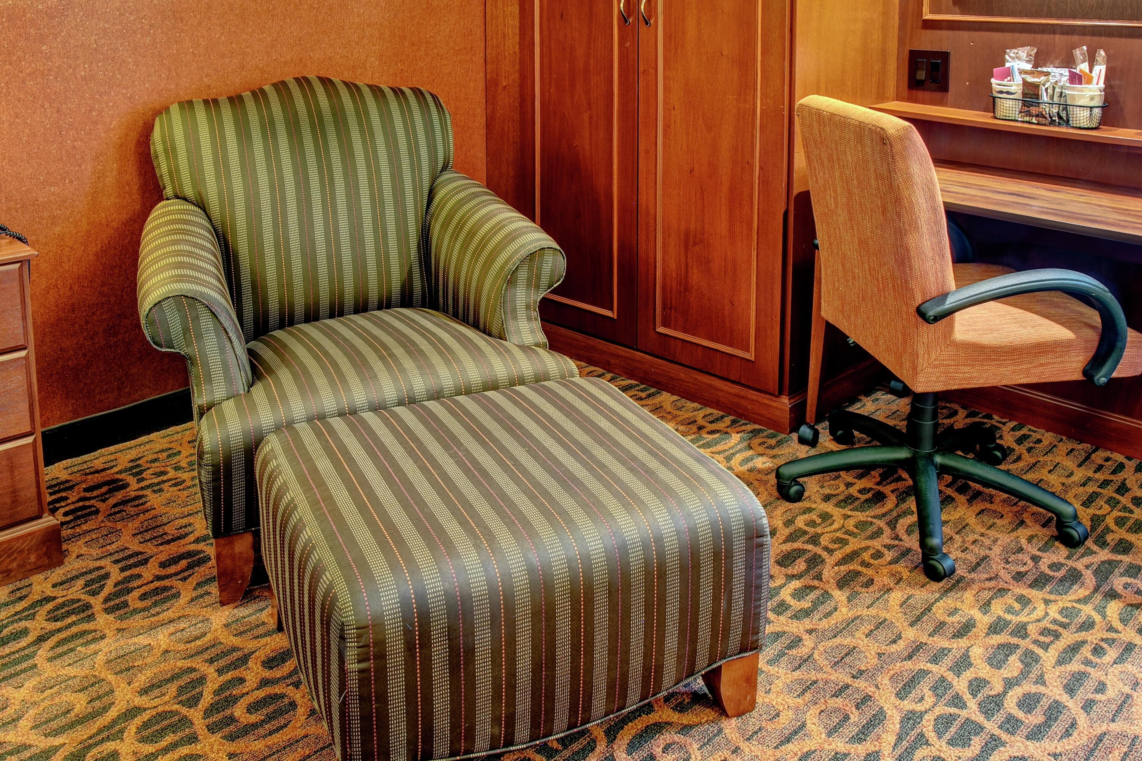 A room with a striped armchair, a matching ottoman, a wooden desk, and a patterned carpet.