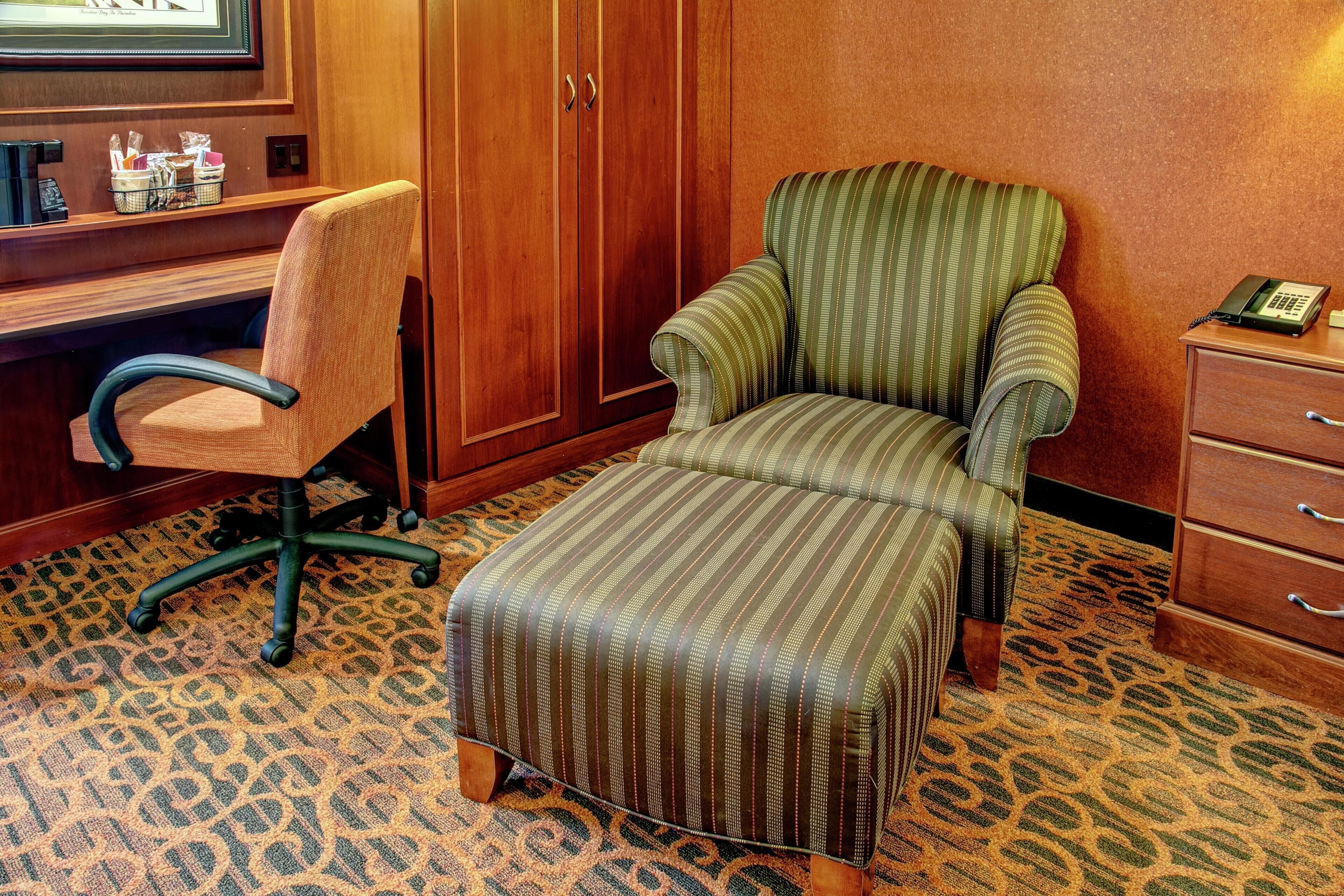 A hotel room with a wooden desk, a striped armchair, a matching ottoman, a telephone, and a patterned carpet.