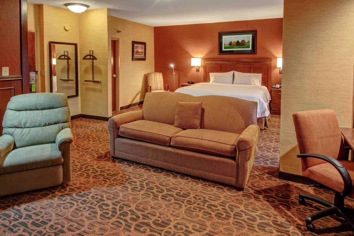 A hotel room with a patterned carpet, a bed, a sofa, a chair, and a desk.