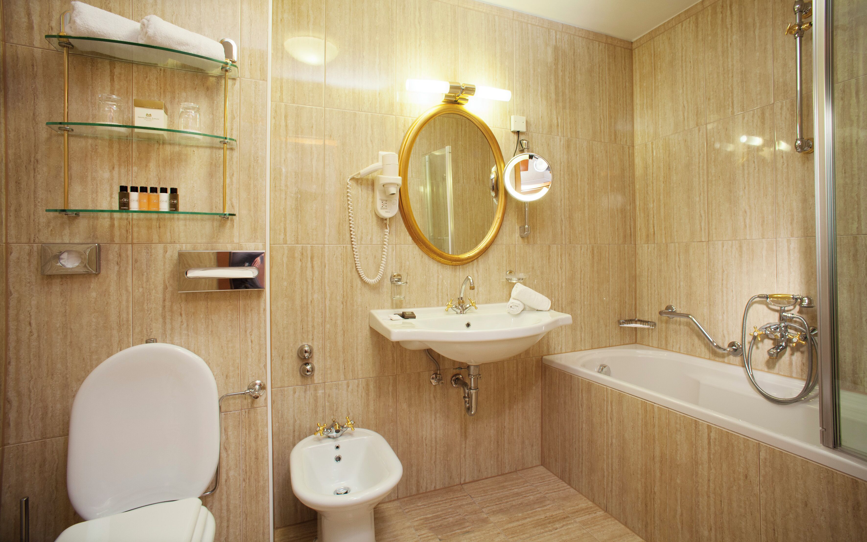 A bathroom with a toilet, bidet, sink, and bathtub. There are shelves with toiletries and a mirror with lights.