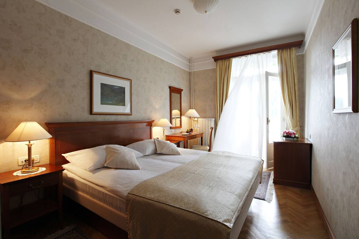 A hotel room with a large bed, two bedside tables with lamps, a nightstand, a wooden headboard, a framed picture on the wall, a mirror, a window with curtains, and a small table with a vase of flowers.