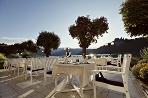 An outdoor dining area with white tables and chairs, a waiter, and a view of the lake and mountains. - Grand Hotel Toplice - Sava Hotels & Resorts (Bled)