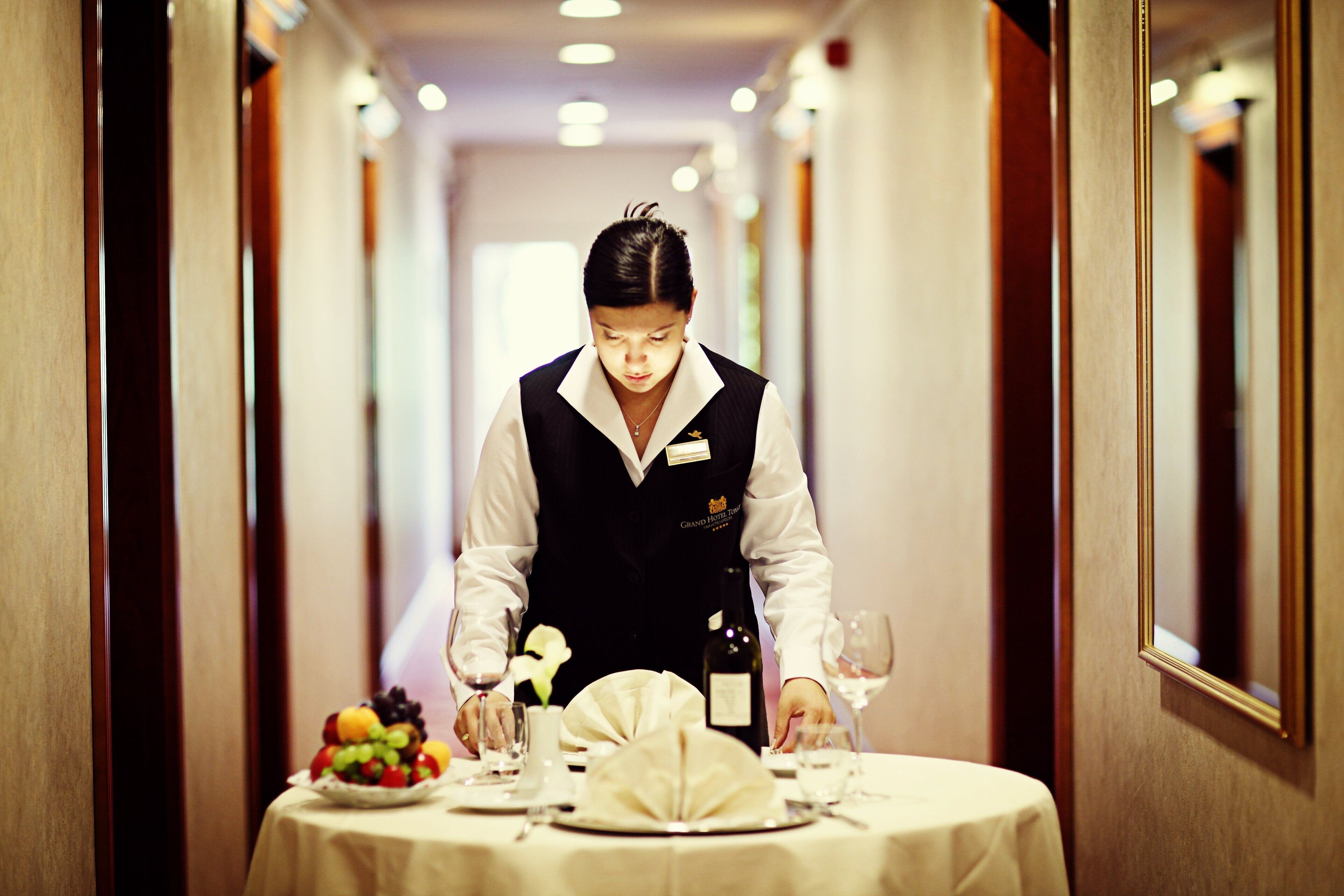 A hotel corridor with a table set for a meal, including a fruit bowl, wine glasses, and a bottle.