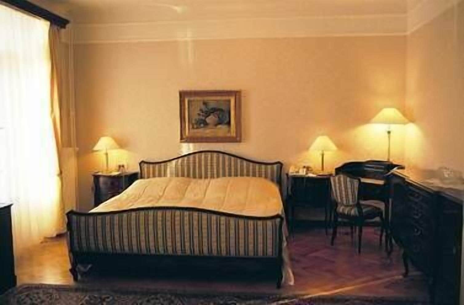 A hotel room with a bed, a desk, two lamps, a chair, and a painting on the wall.