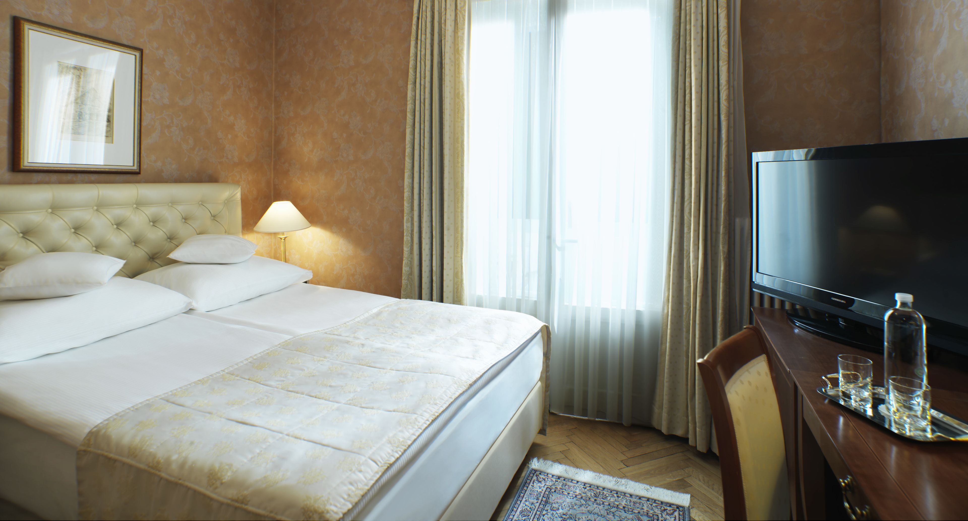 A hotel room with a large bed, a television, a bedside table with glasses and a bottle, and a framed picture on the wall.