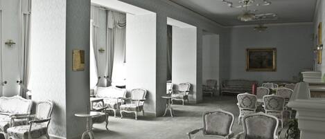 Ein geräumiges Zimmer mit aufwendigen Stühlen und Tischen, großen Fenstern mit Vorhängen und einer Kristallleuchter.