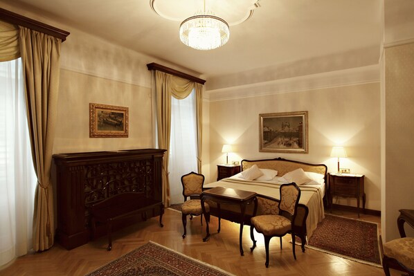 A hotel room with a large bed, two bedside tables with lamps, a wooden dresser, a desk with a chair, and a large rug.
