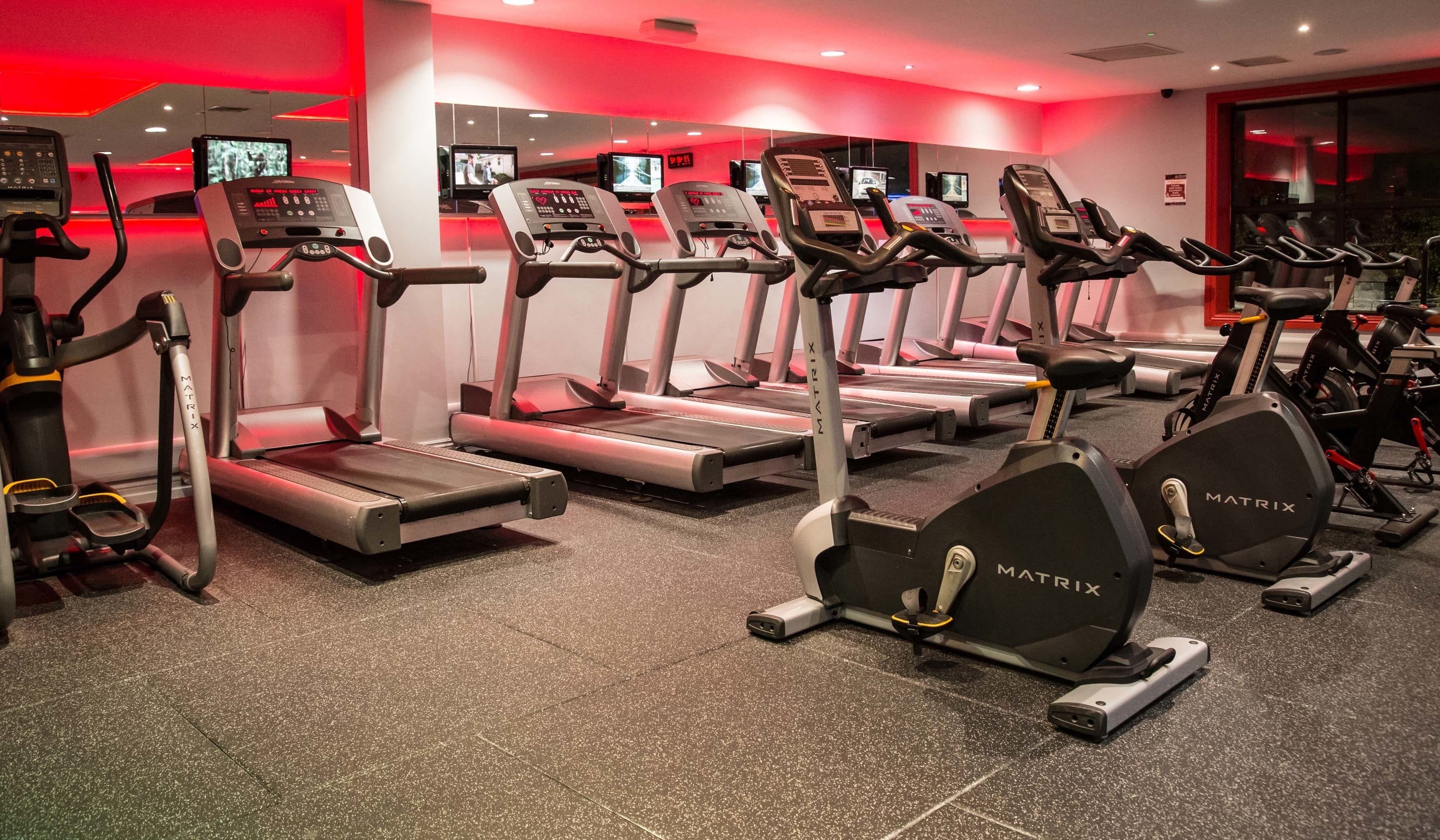Un gimnasio con múltiples cintas de correr y máquinas elípticas, equipadas con pantallas digitales y monitores de frecuencia cardíaca.