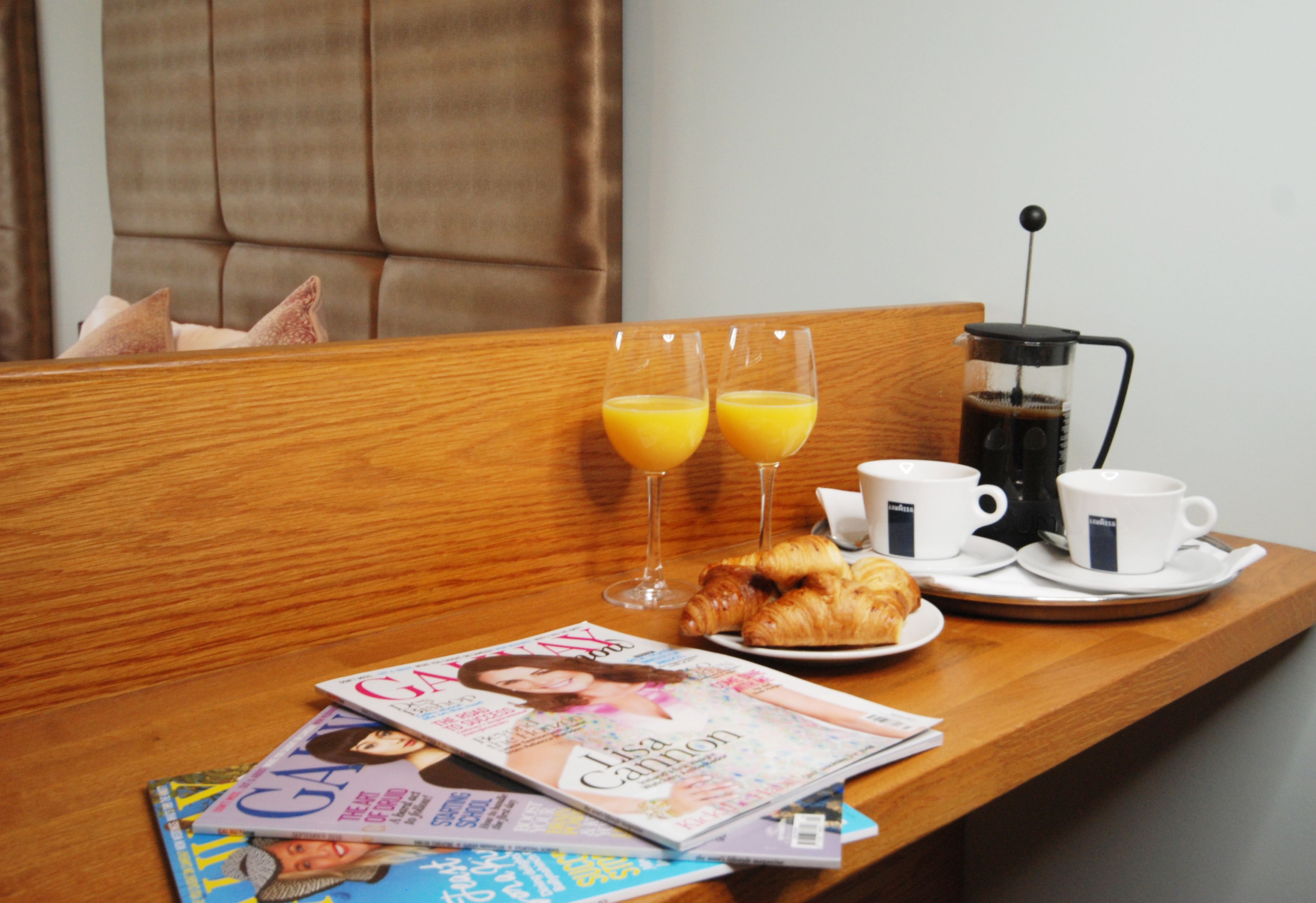 A wooden table with breakfast items including croissants, two glasses of orange juice, a coffee maker, and a magazine.