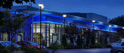 A modern hotel with a glass facade, parking lot, and street lamps.