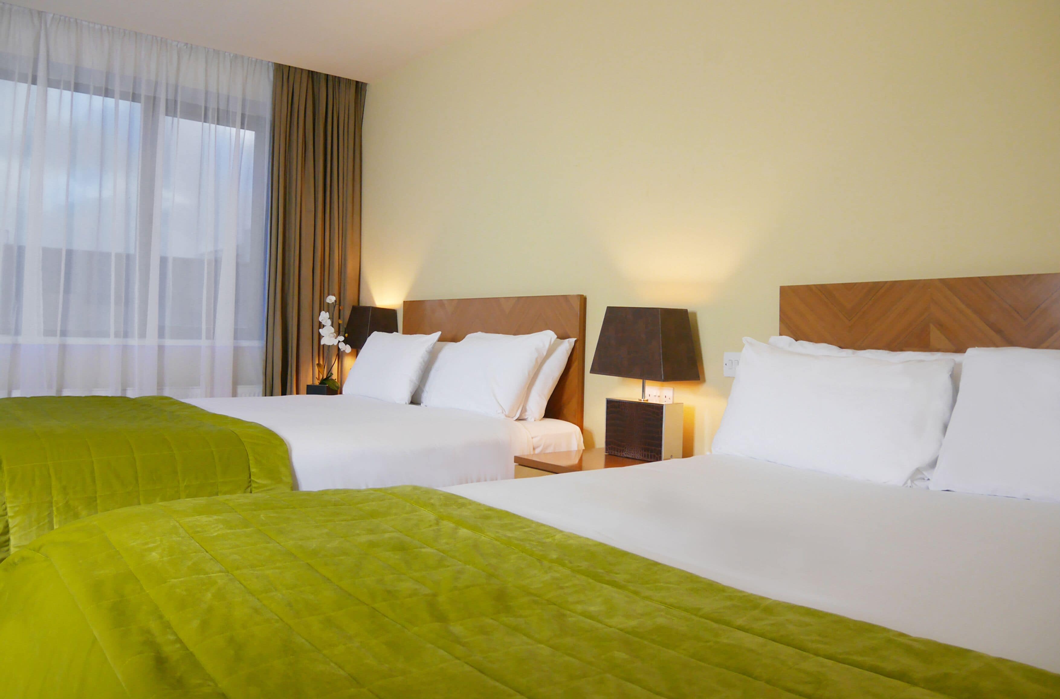 A hotel room with two beds, each with white linens and green bedspreads. There are two bedside tables with lamps, and a large window with sheer curtains.