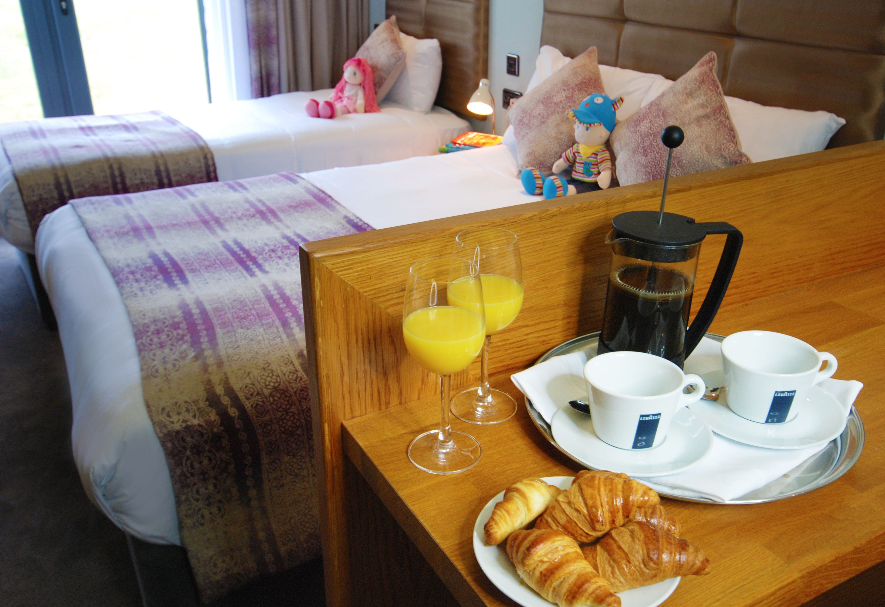 Een hotelkamer met twee bedden, een ontbijtserver met croissants, sap, koffie en bekers, en uitzicht op de kamer.