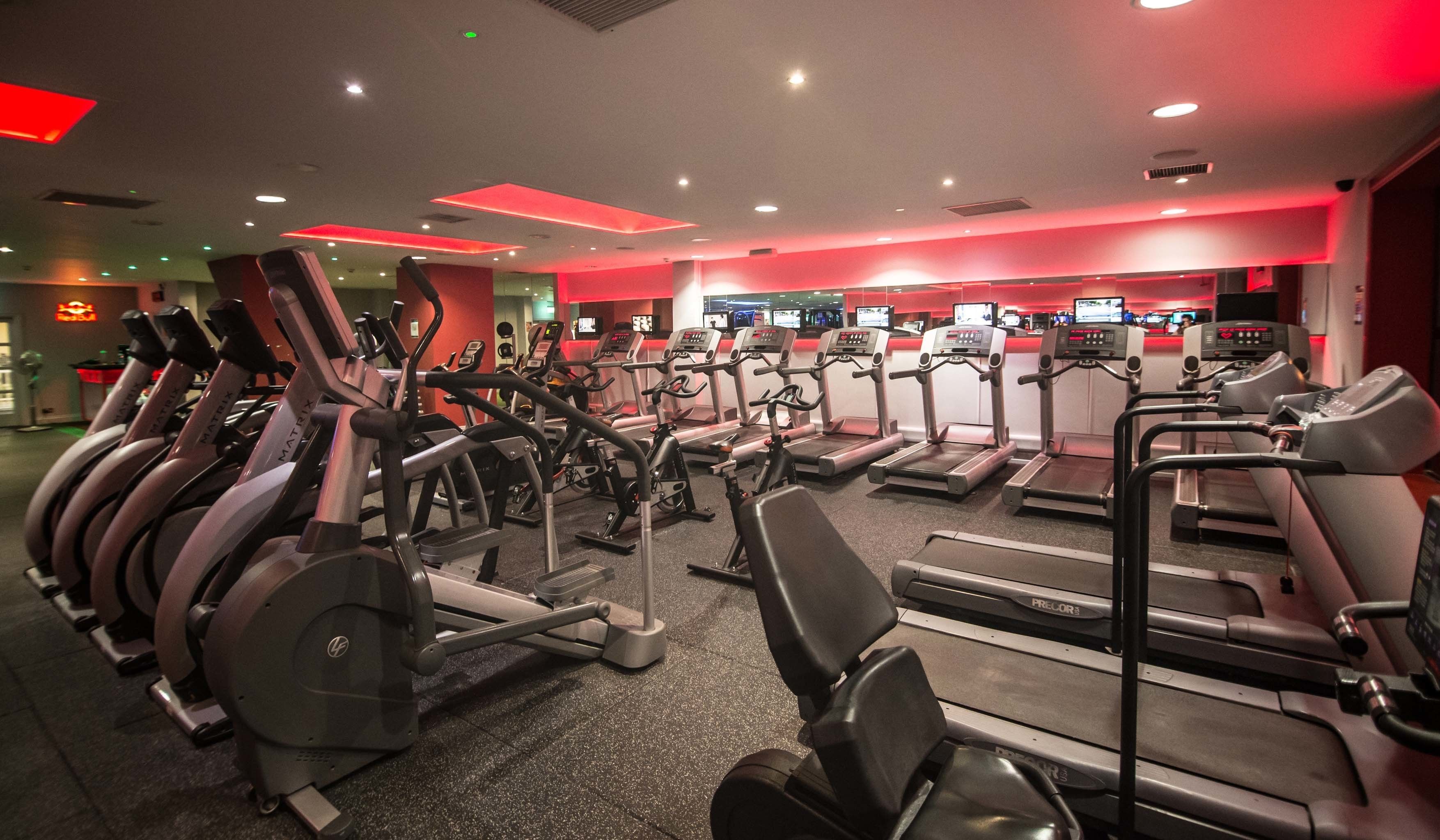 Une salle de sport équipée de plusieurs tapis de course et de plusieurs appareils elliptiques, avec un plafond éclairé en rouge et un panneau Red Bull bien visible à l’arrière-plan.