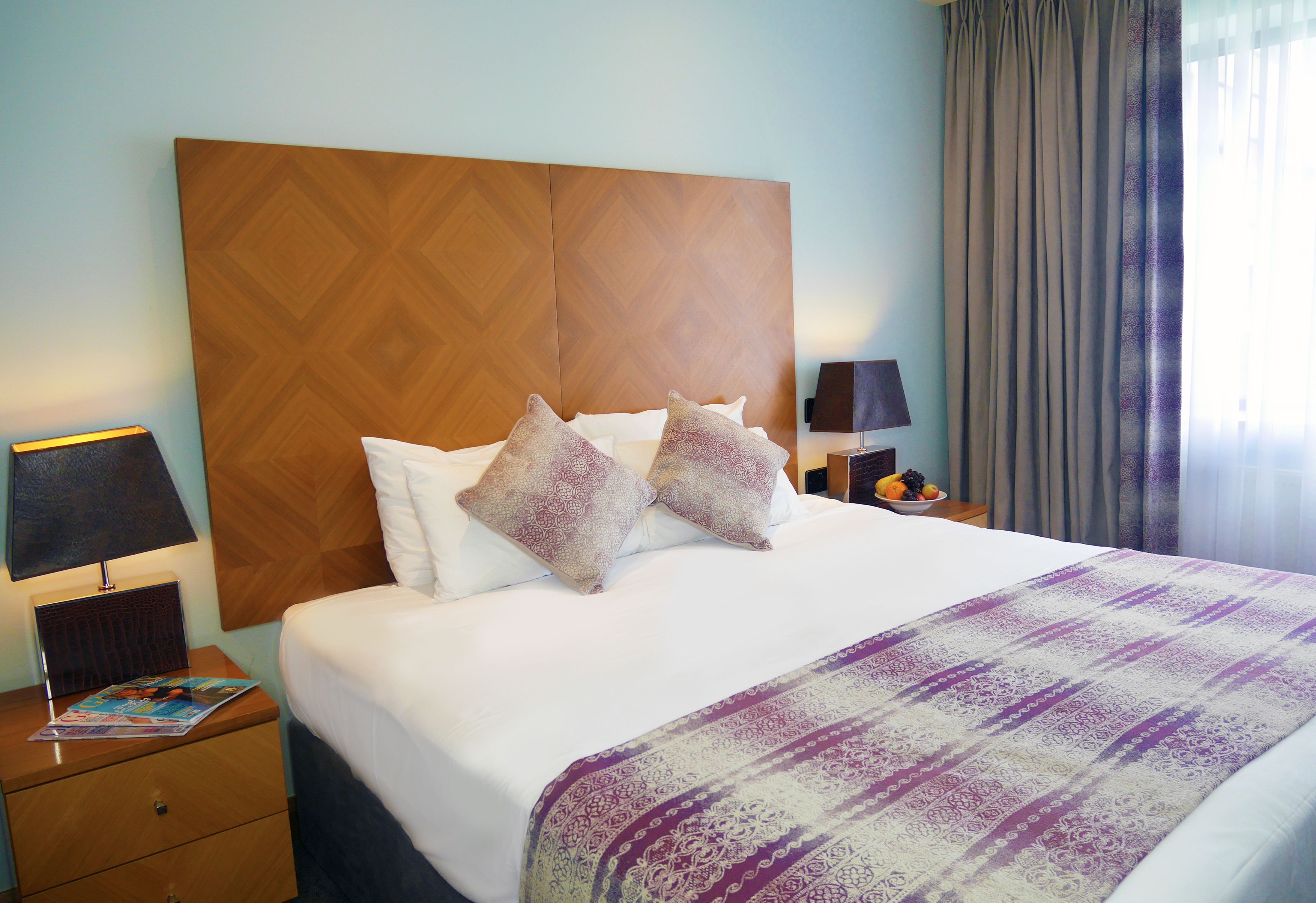 A hotel room with a large bed, two nightstands, a wooden headboard, and a view of the outdoors through sheer curtains.