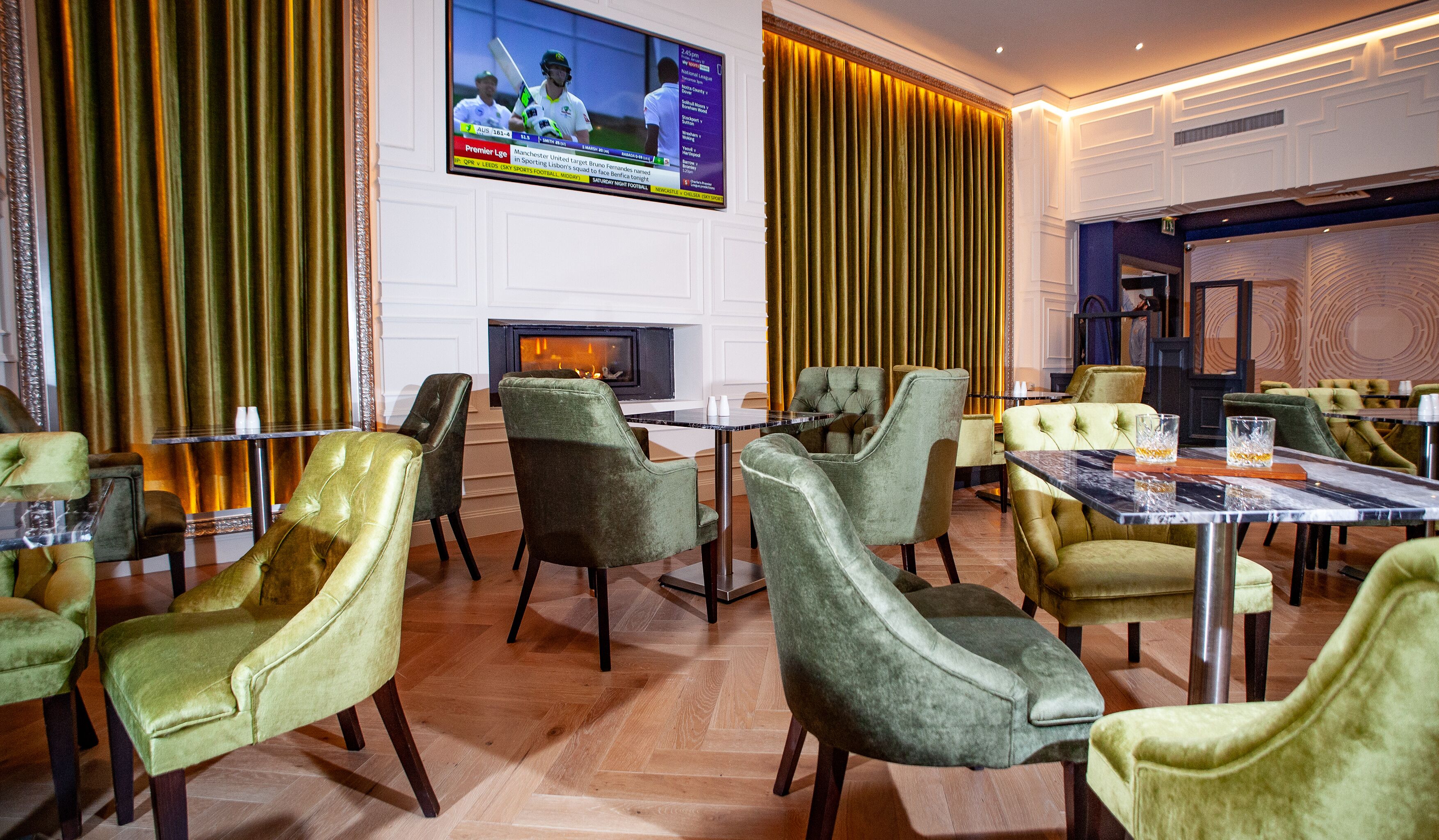 A hotel lobby with green upholstered chairs, a fireplace, and a television displaying a sports event.
