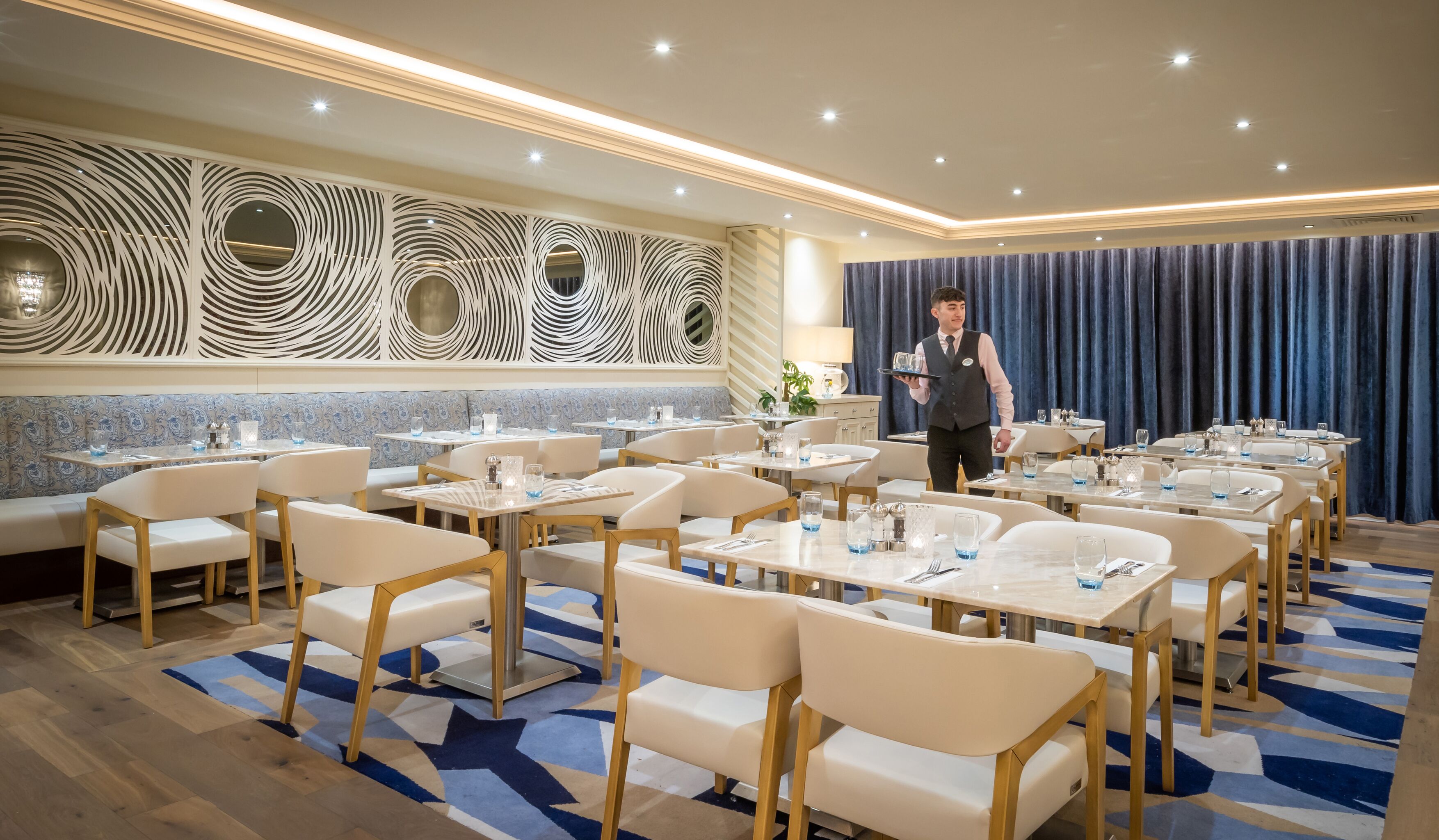 A hotel dining area with multiple tables and chairs, a waiter serving, and a decorative wall with circular patterns.