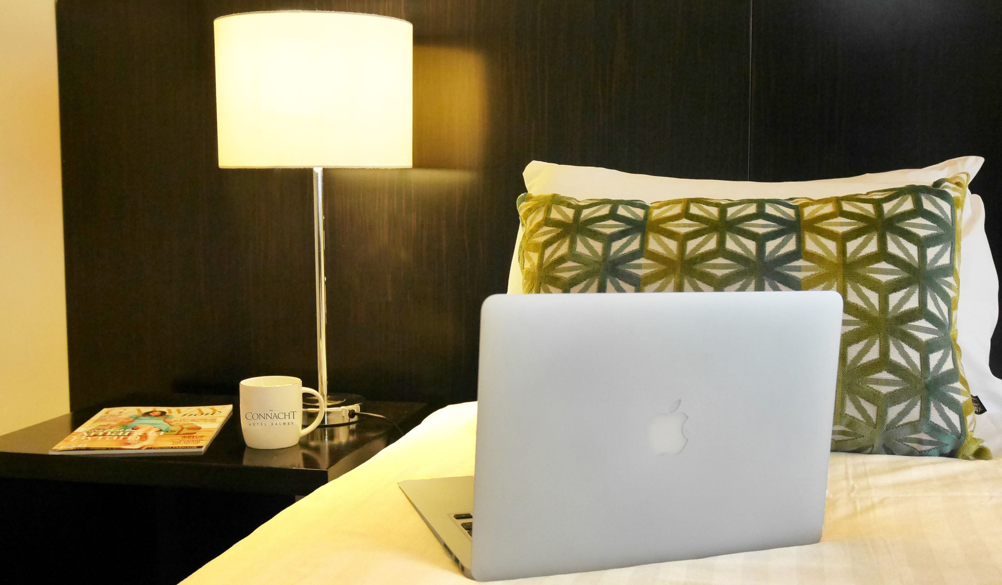 Hotellihuone, jossa on sänky, kannettava tietokone, lehti, muki ja lamppu.