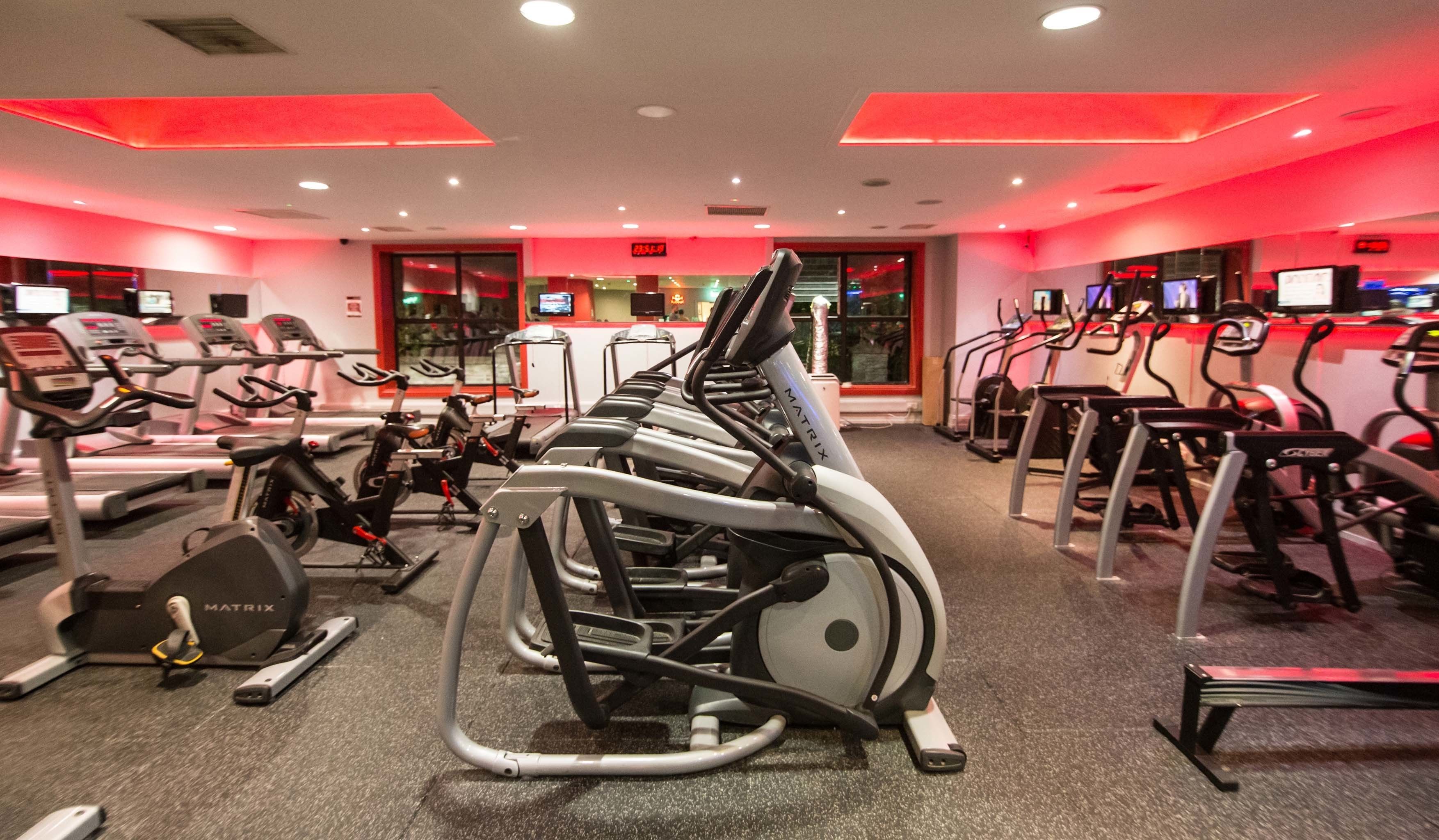 A gym with multiple treadmills and stationary bikes, red lighting, and a digital display on the wall.