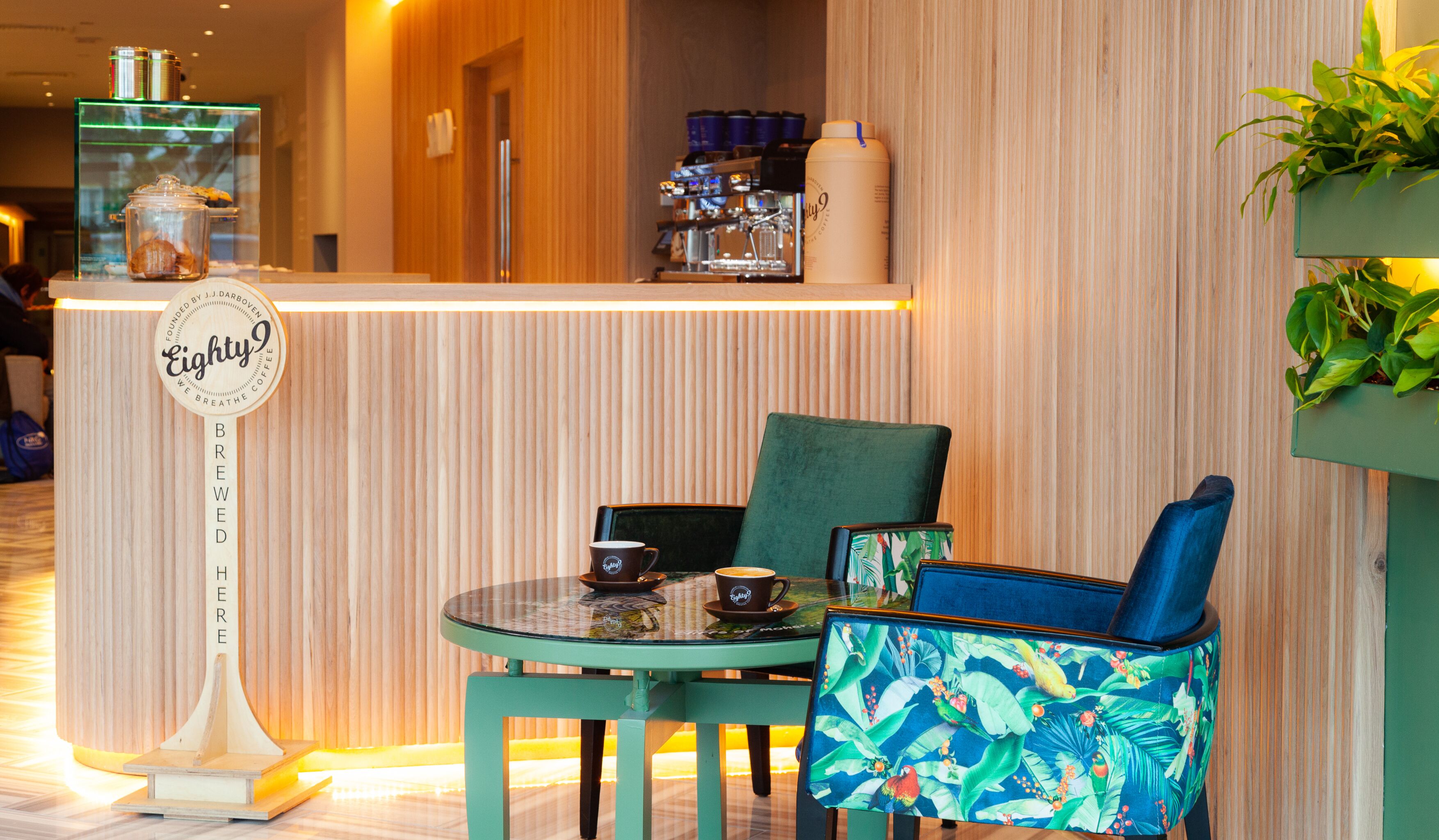 Un café con interior de madera, una barra con una máquina de café y dos sillas con estampado tropical.
