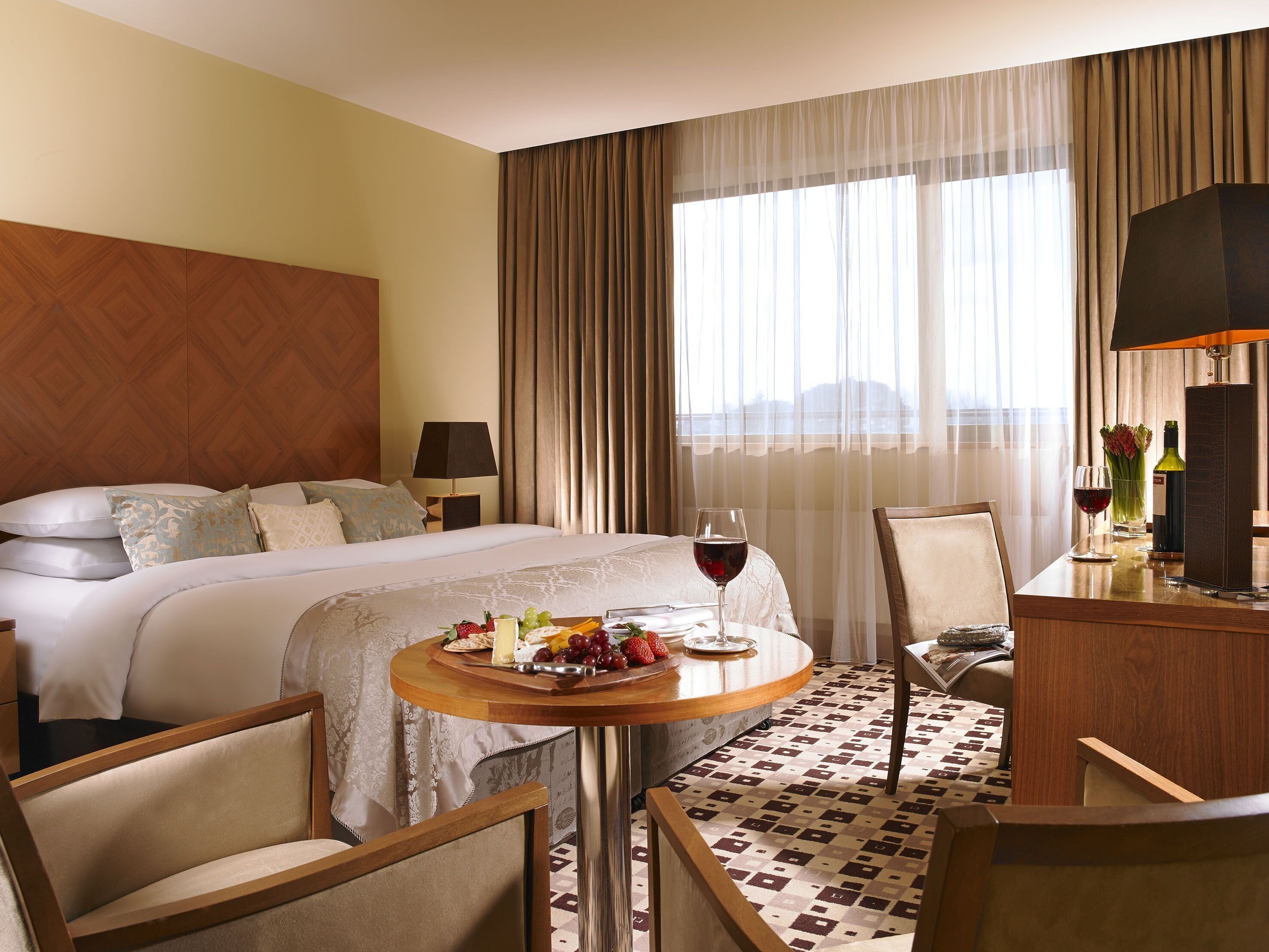 Une chambre d’hôtel avec un lit, une table à manger avec des fruits et du vin, un bureau avec une lampe et une vue sur la ville grâce à de grandes fenêtres.