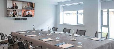 A conference room with a long table, chairs, a large screen for video conferencing, and large windows.