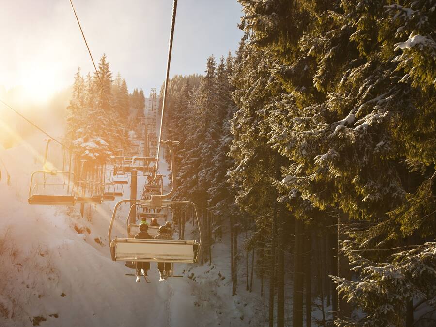 A ski lift with passengers ascending a snowy slope, surrounded by evergreen trees.