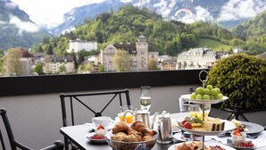 Ein Frühstückstisch mit Croissants, Käse und Obst, vor einer Berglandschaft und einer Stadt.