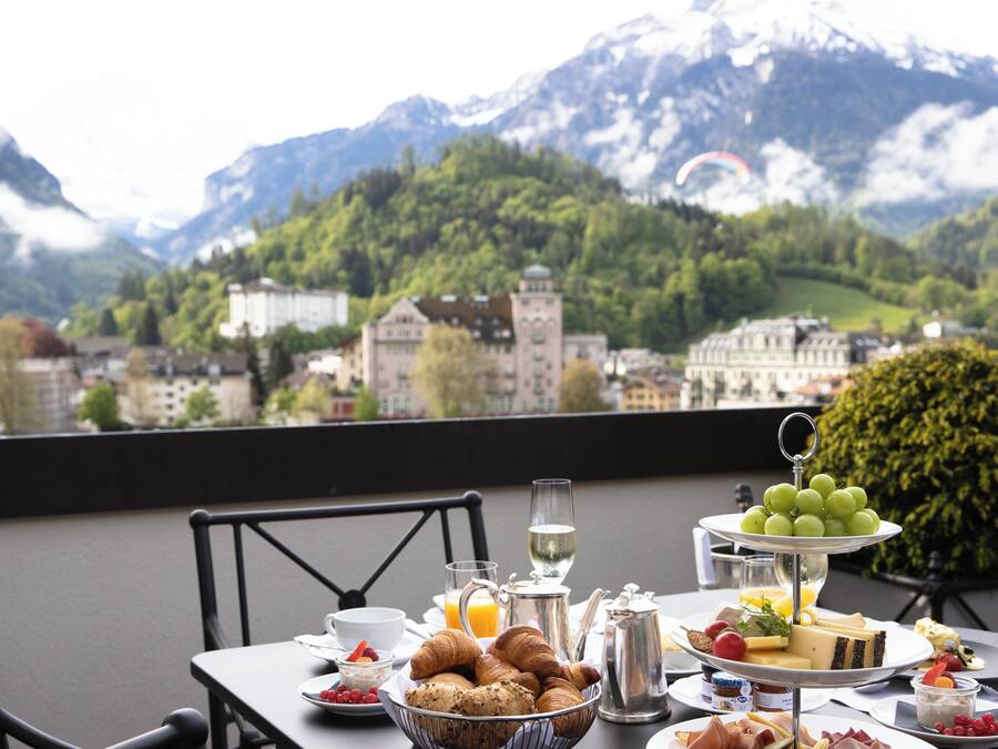 A breakfast table with croissants, cheese, and fruit, set against a backdrop of mountains and a town.