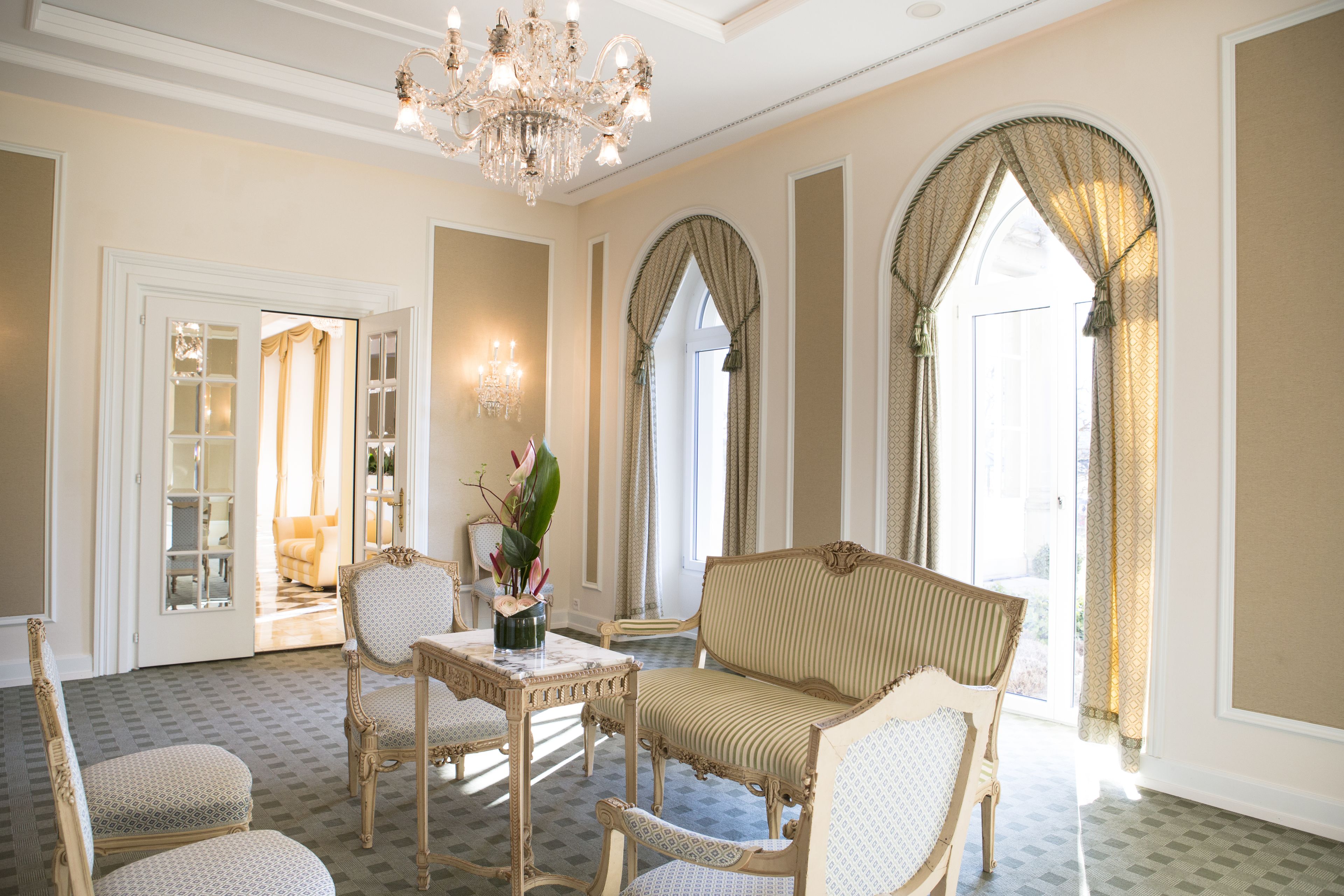 A room with a chandelier, a sofa, and a table with chairs. There are windows with curtains and a door leading to another room.