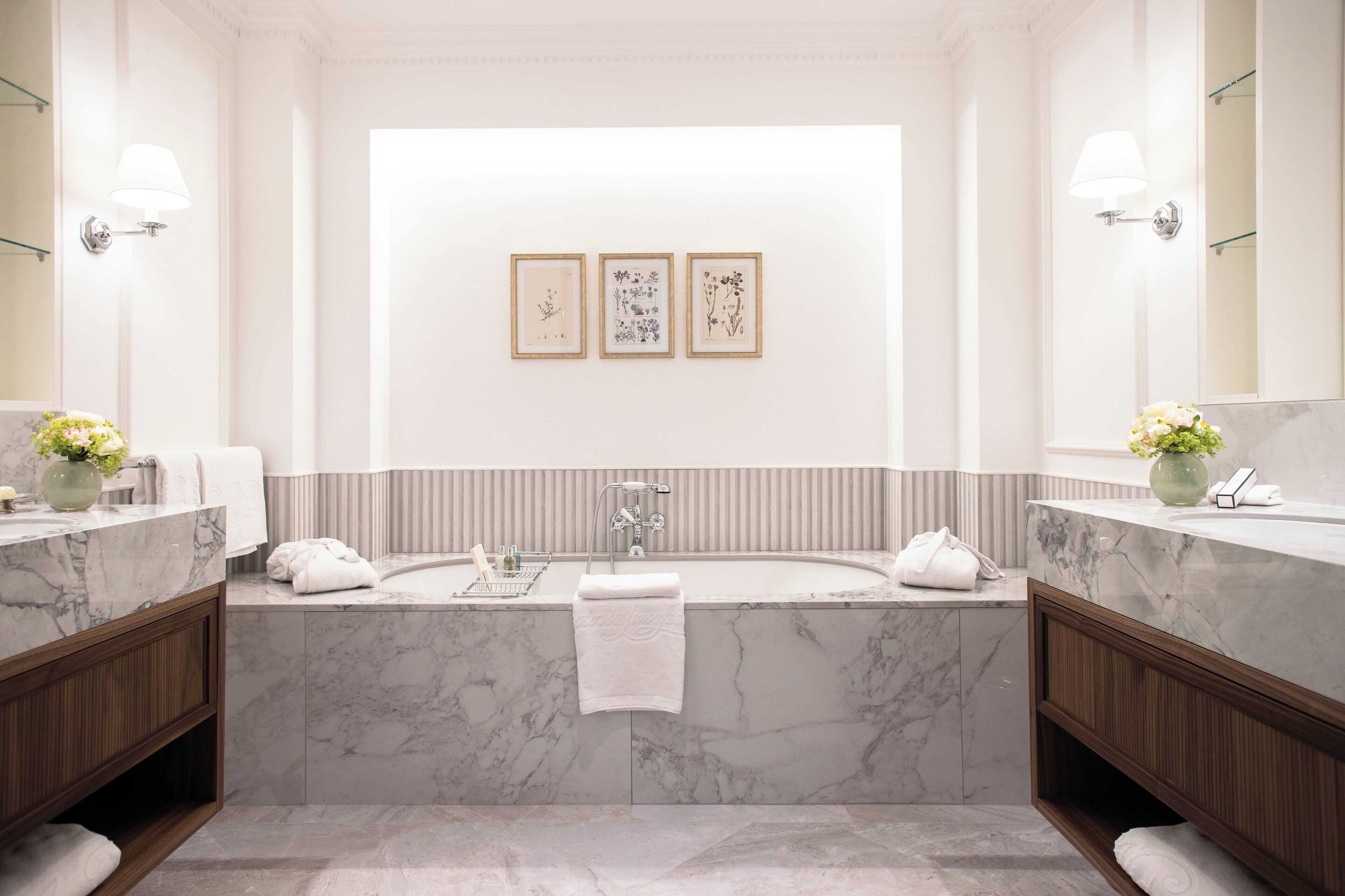 Une salle de bain moderne avec une grande baignoire, deux vasques, des plans de travail en marbre et des œuvres d’art botanique encadrées accrochées au mur.
