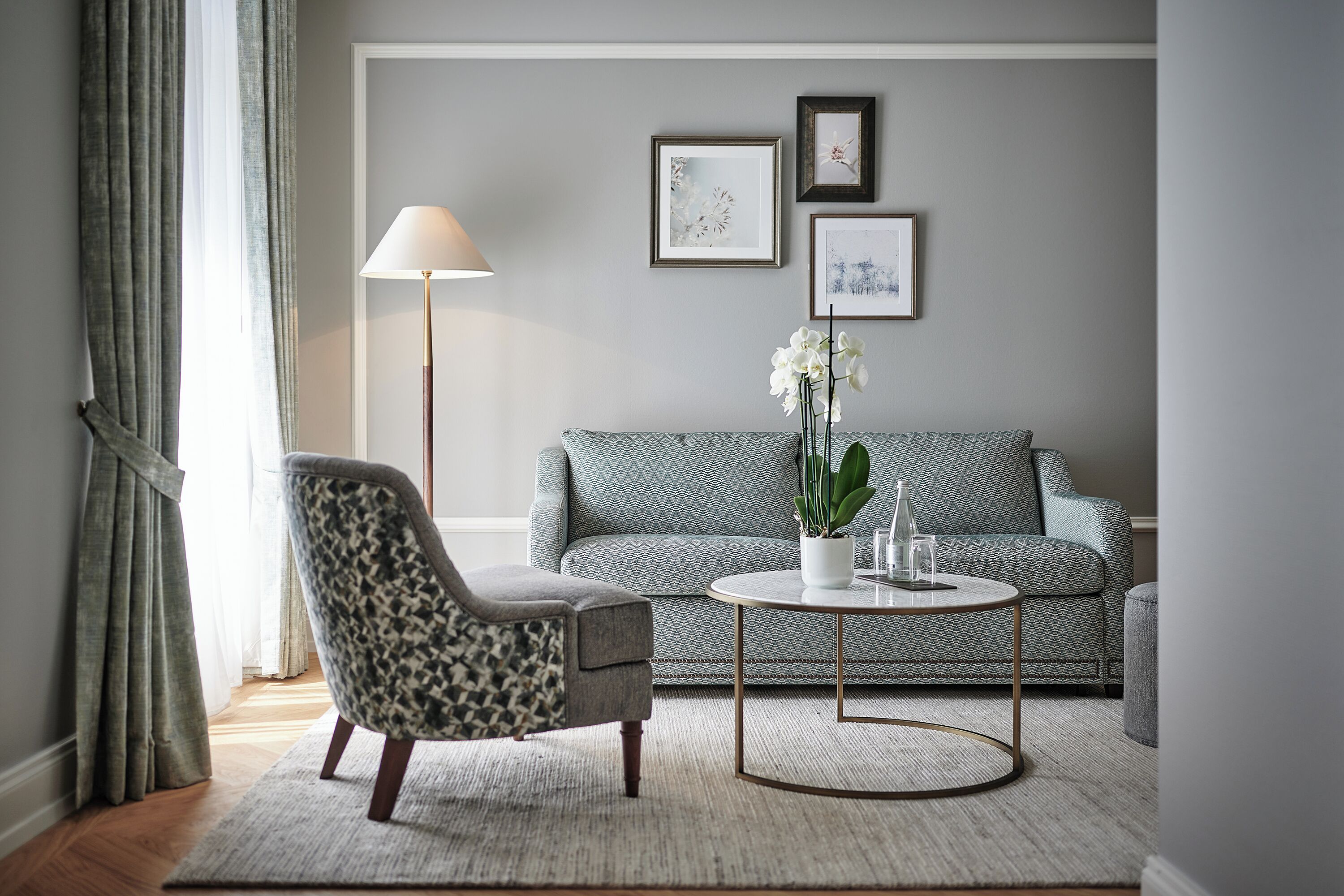 A living room with a patterned sofa, a patterned armchair, a round coffee table with a plant, and a floor lamp.