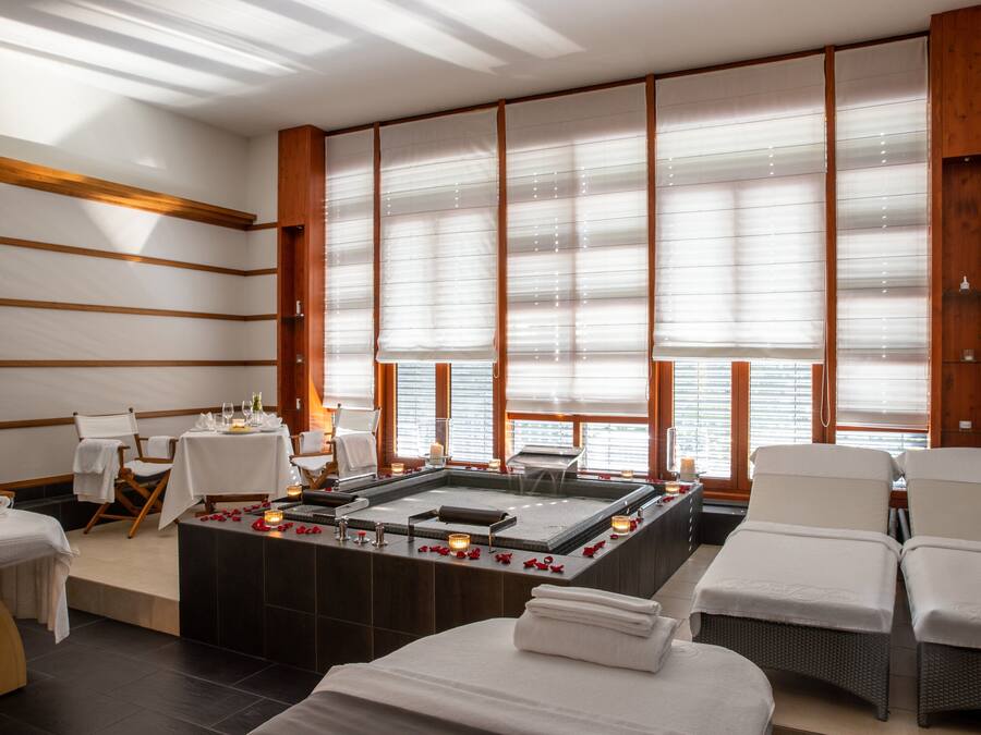 Een spa ruimte met een massagetafel, een massagestoel, een klein tafeltje met kaarsen en een groot raam met luxaflex.