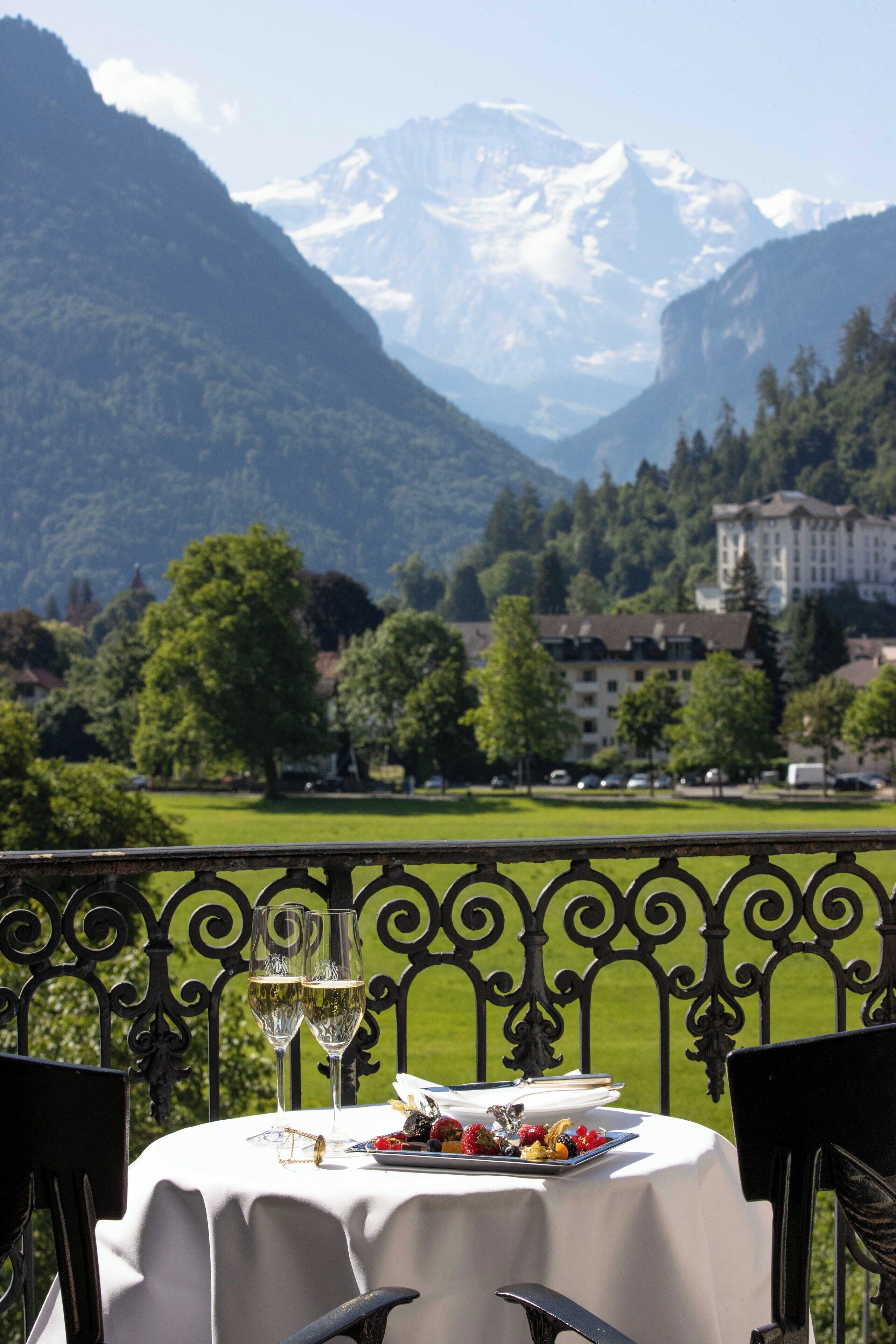 A table set with a meal and glasses of wine, overlooking a mountainous landscape with a building in the background.