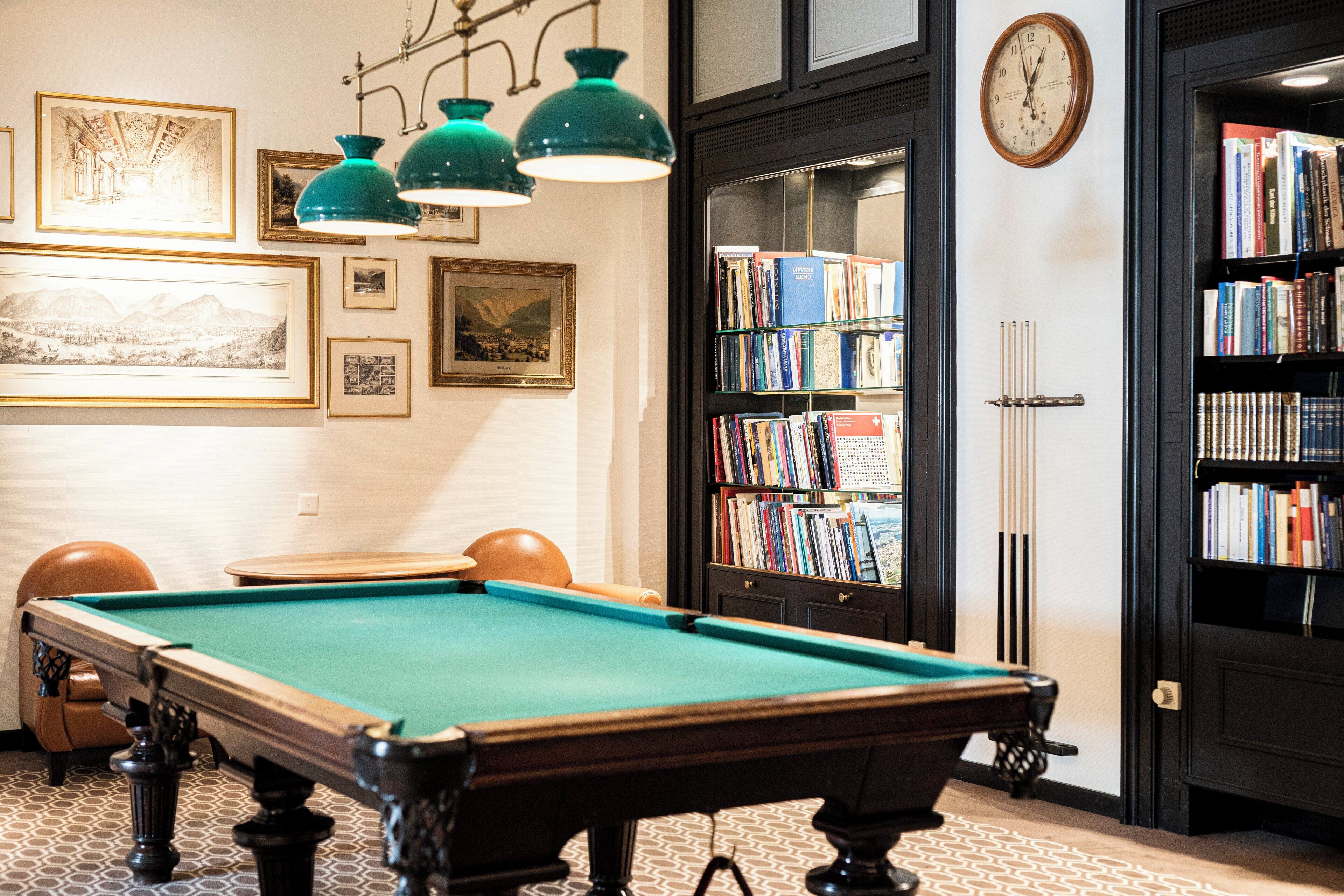 Ein Billardtisch mit grüner Spielbahn, umgeben von gerahmten Bildern an den Wänden, einem Bücherregal voller Bücher und einer Wanduhr.
