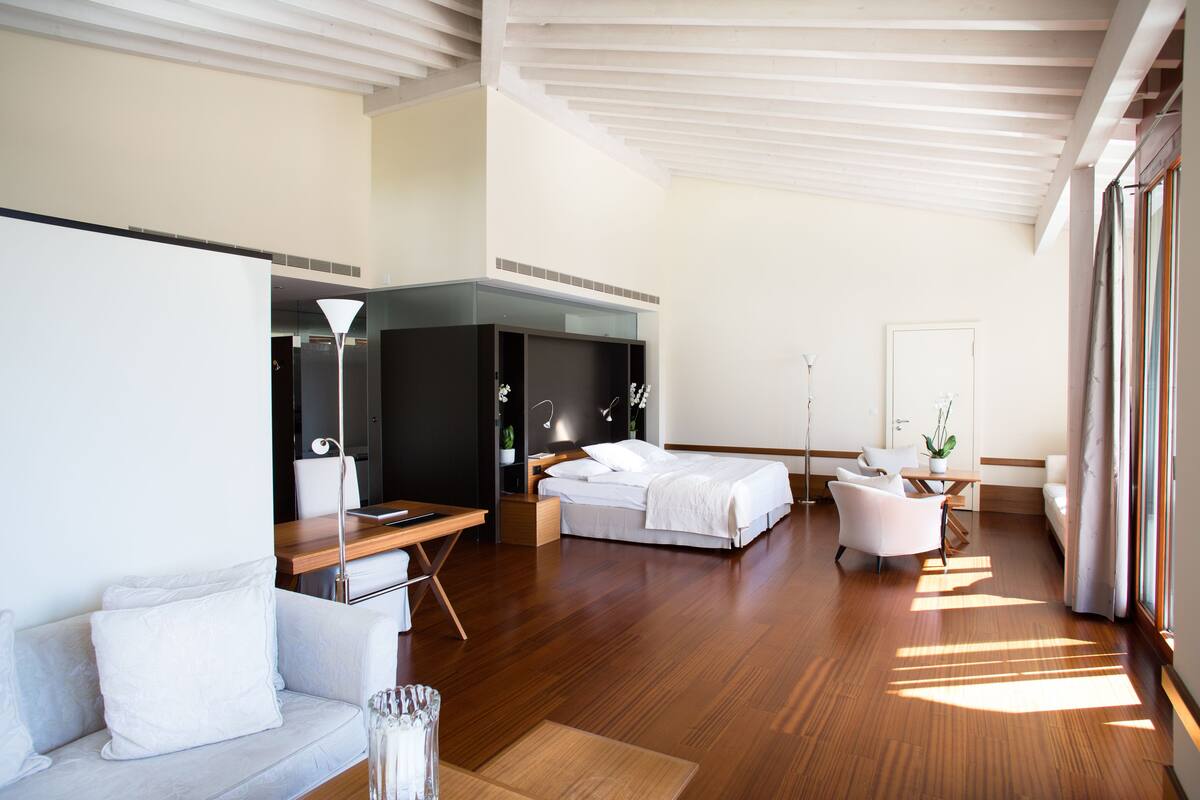 Una habitación de hotel moderna con suelo de madera, una cama, un sofá, un escritorio y un ventanal grande.