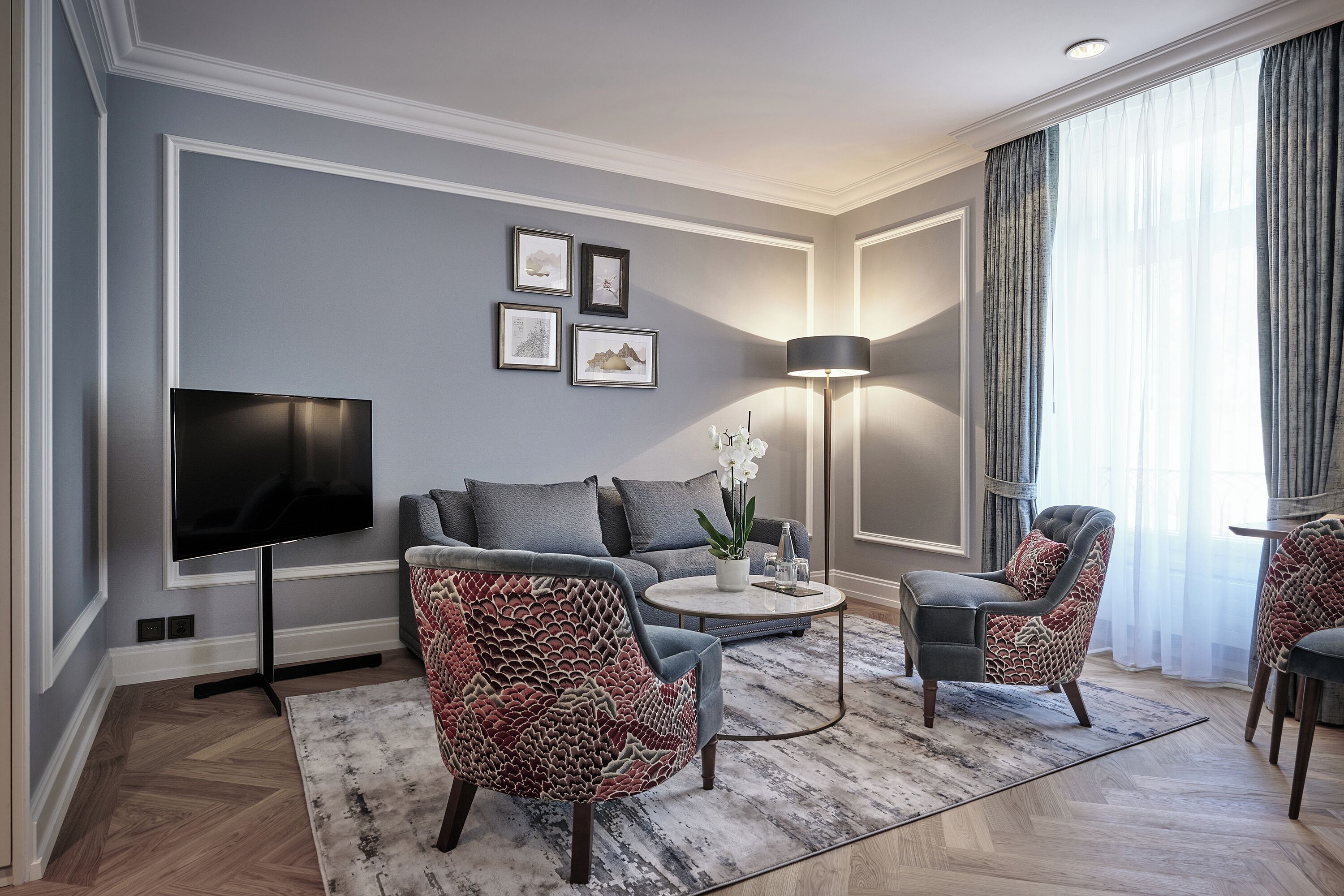 Una sala de estar moderna con televisor de pantalla plana, un sofá gris, sillones estampados, una mesa de centro y una l ámpara de pie.