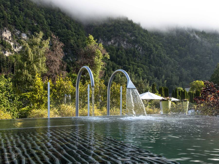 Una piscina con chorros de agua, rodeada de abundante vegetación y montañas al fondo.
