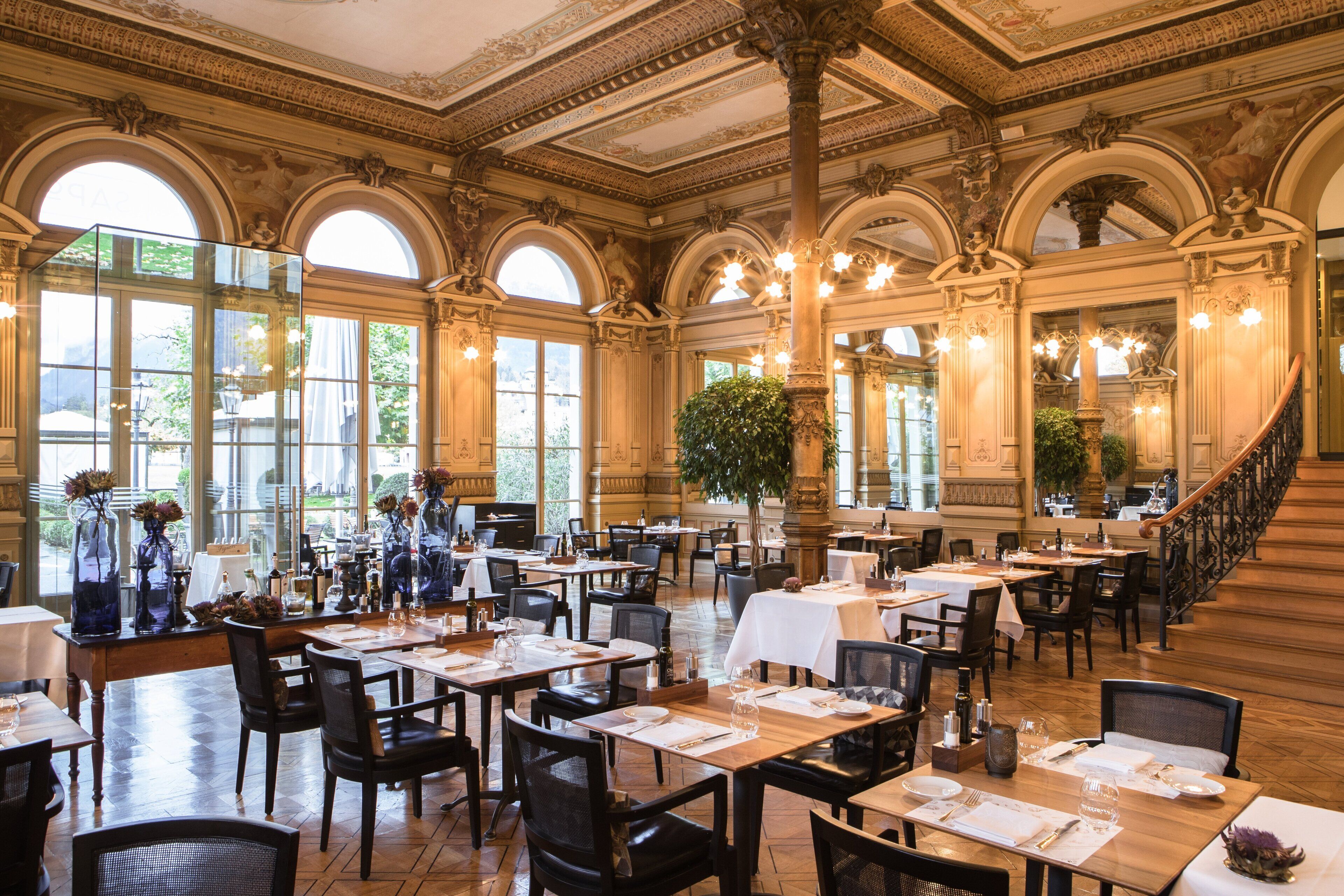 Un comedor amplio con varias mesas y sillas, ventanas grandes y detalles ornamentales en el techo.