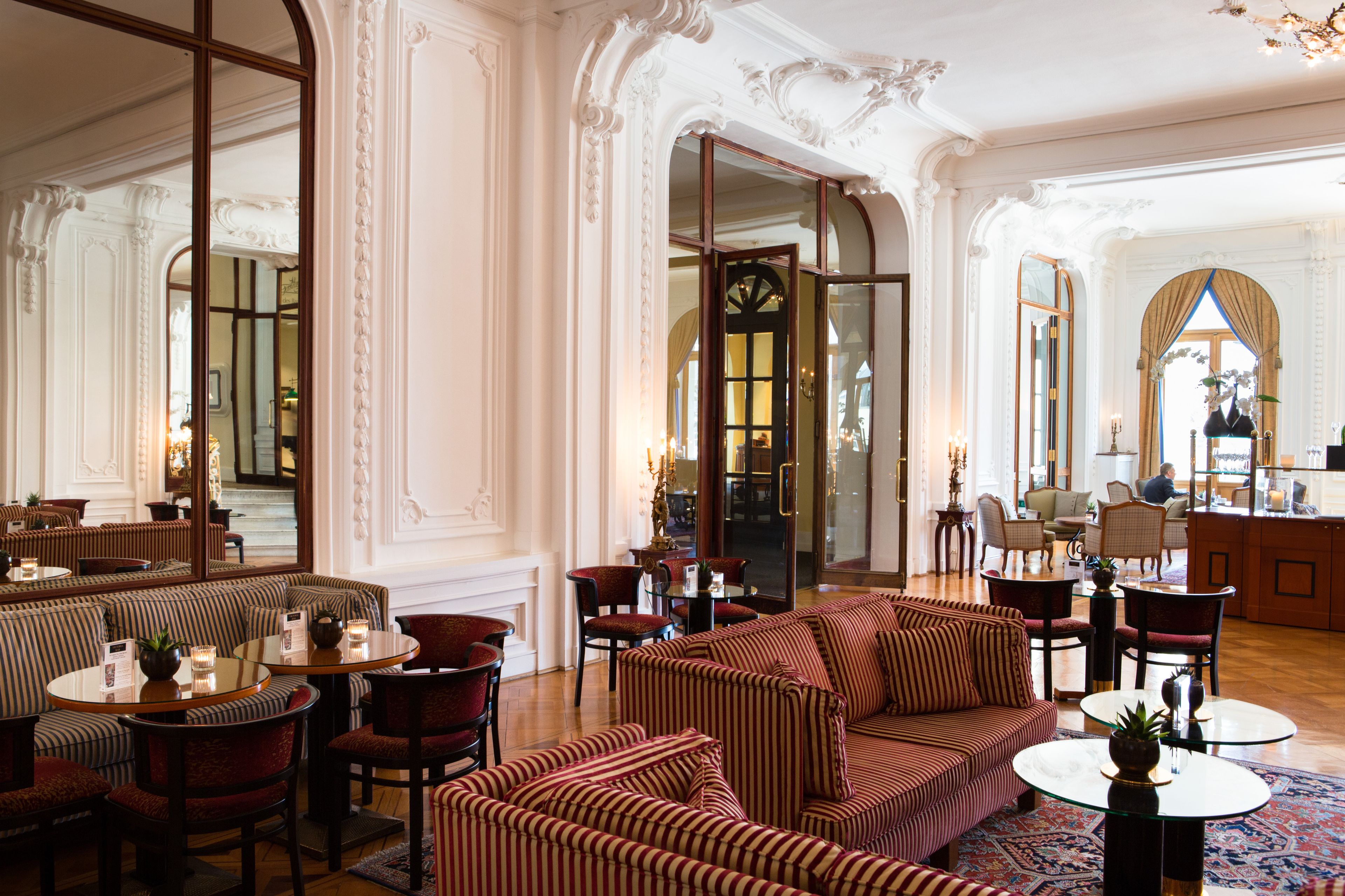 Un lobby de hotel lujoso con paredes blancas ornamentadas, una escalera monumental y un área de estar con sofás y sillones a rayas.