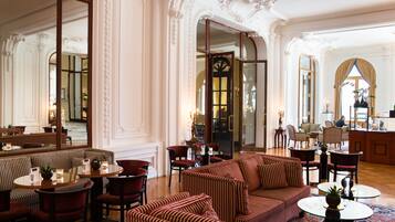 Un lobby de hotel lujoso con paredes blancas ornamentadas, una escalera monumental y un área de estar con sofás y sillones a rayas.